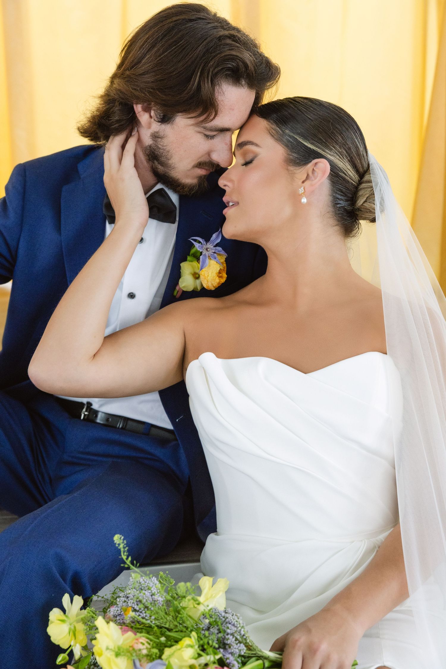 Romantic newlyweds share an intimate moment in formal attire with the bride holding a yellow bouquet against golden drapes.