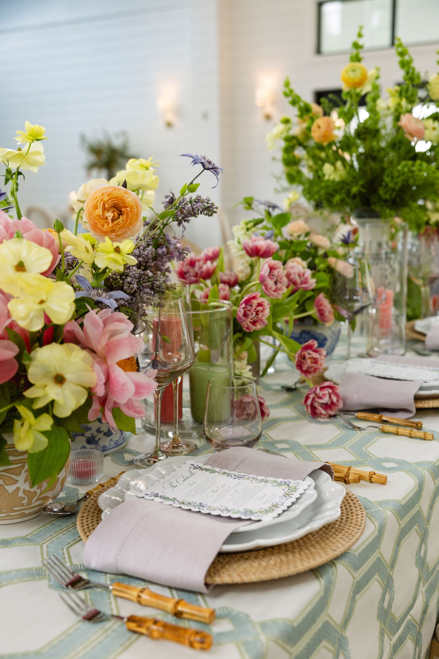 Elegant table setting with colorful spring flowers in glass vases, woven placemats, and bamboo flatware on patterned tablecloth.