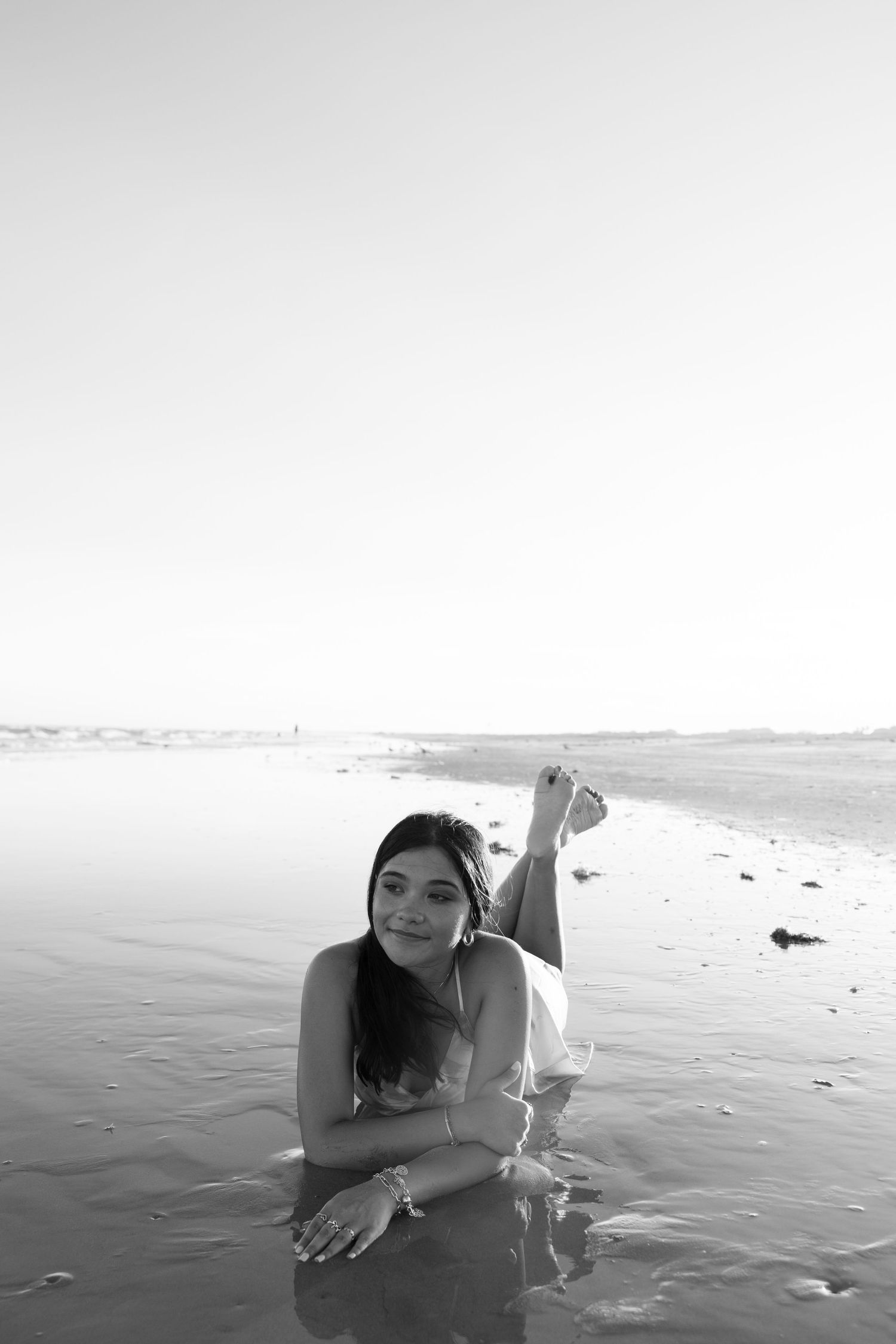 A person lies on a wet beach at the ocean's edge in a black and white artistic photograph.