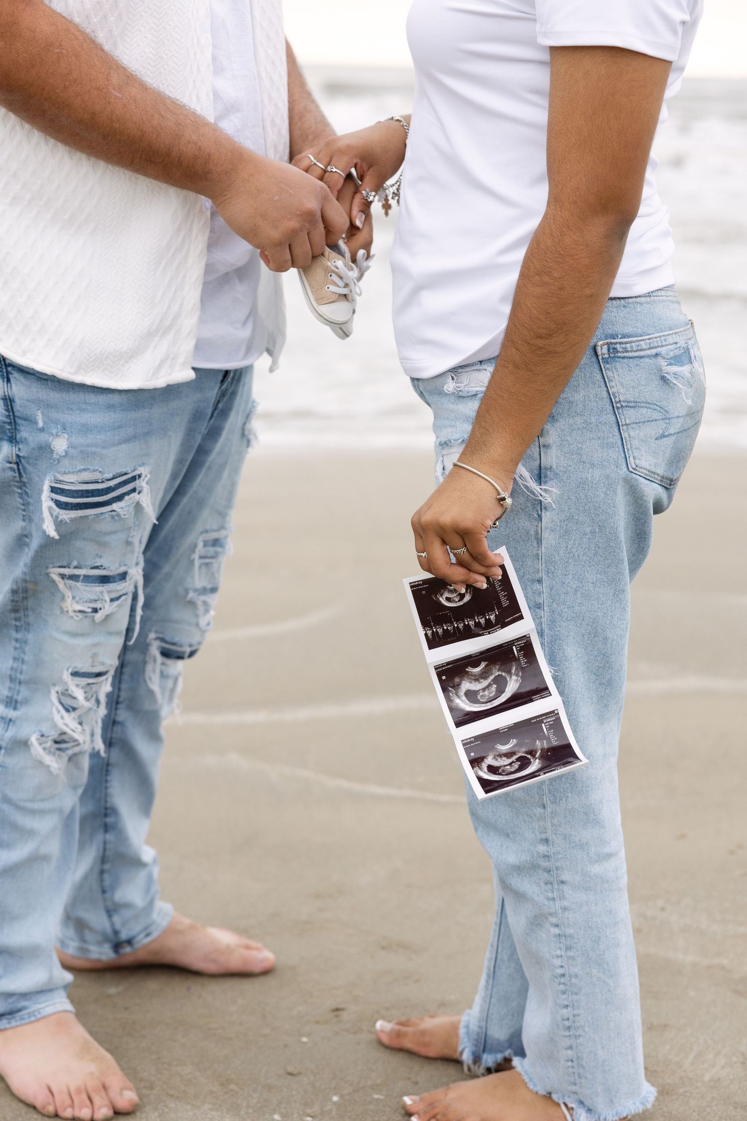 Two people in white shirts and ripped jeans stand barefoot on sand while holding ultrasound photos.