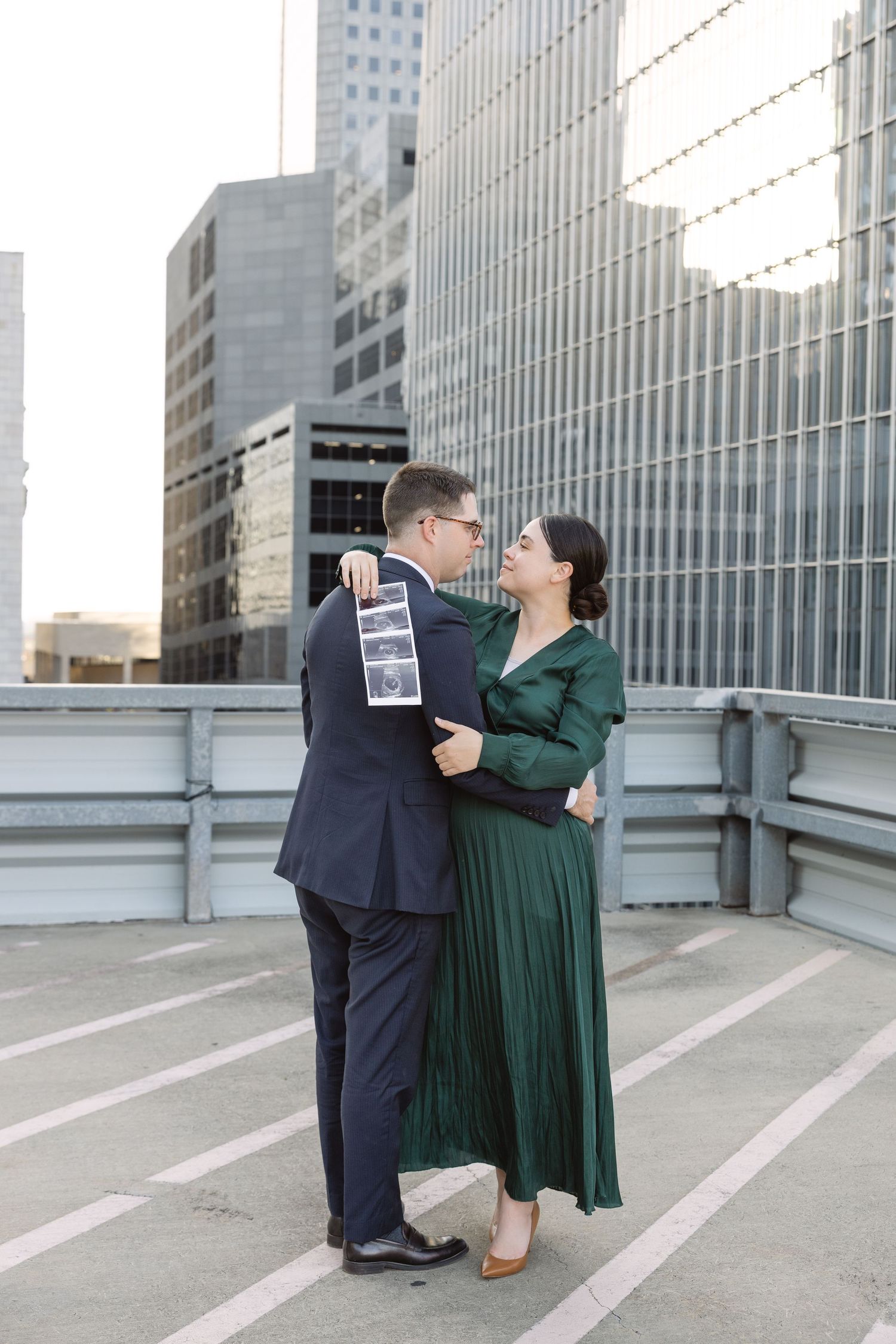 A couple in formal attire share a romantic moment on a city rooftop with modern glass buildings in the background.