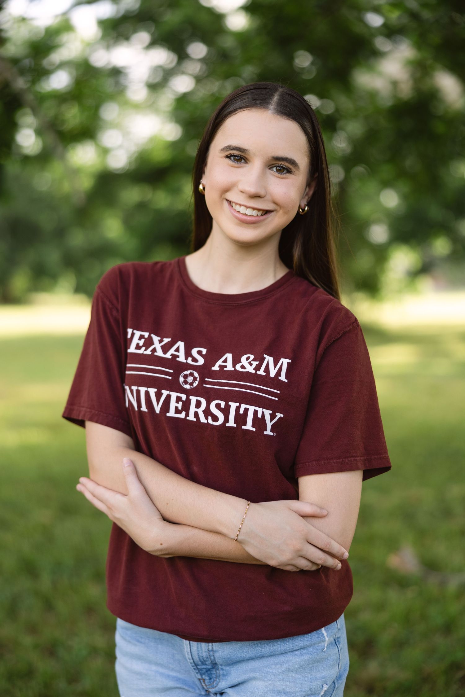 Student wearing maroon Texas A&M University t-shirt poses outdoors with crossed arms against blurred green background.