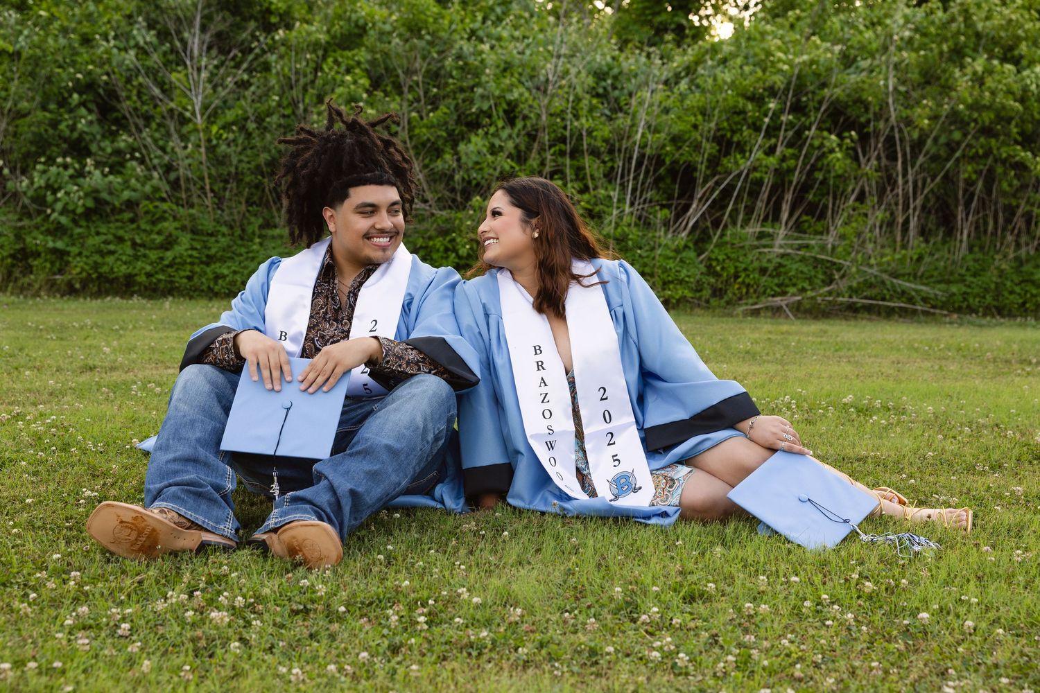 Two graduates in caps and gowns sit together on grass while celebrating commencement on a sunny spring day.
