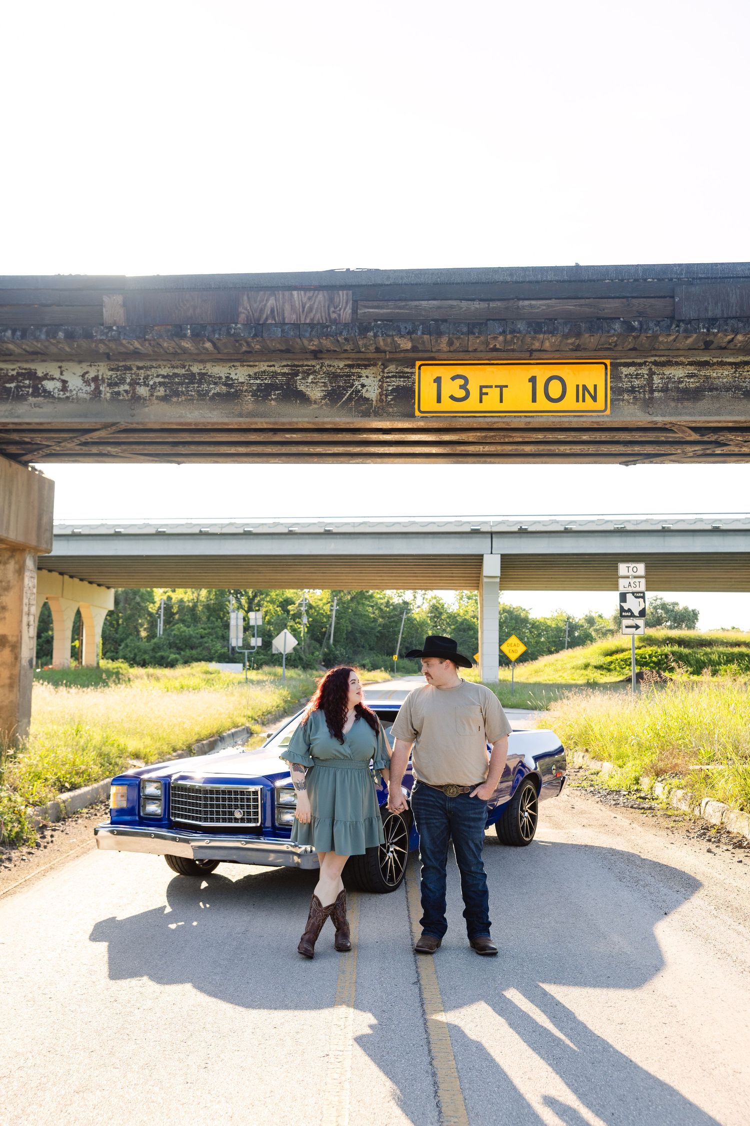 A couple stands beneath a bridge with a 13 ft 10 in clearance sign, posing next to a classic blue car on a rural road.