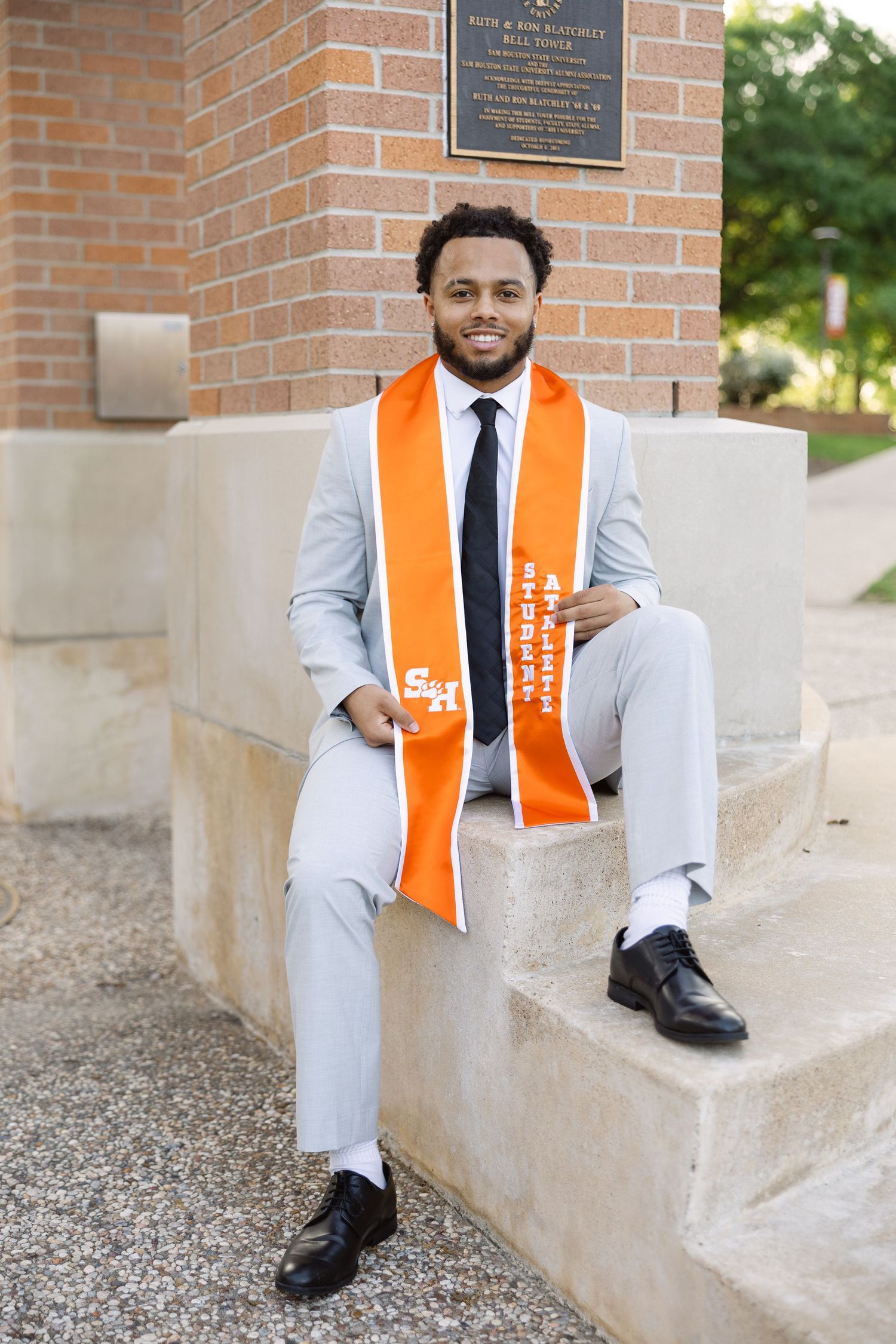 Graduate in light grey suit and orange graduation stole sits on concrete steps near brick wall.