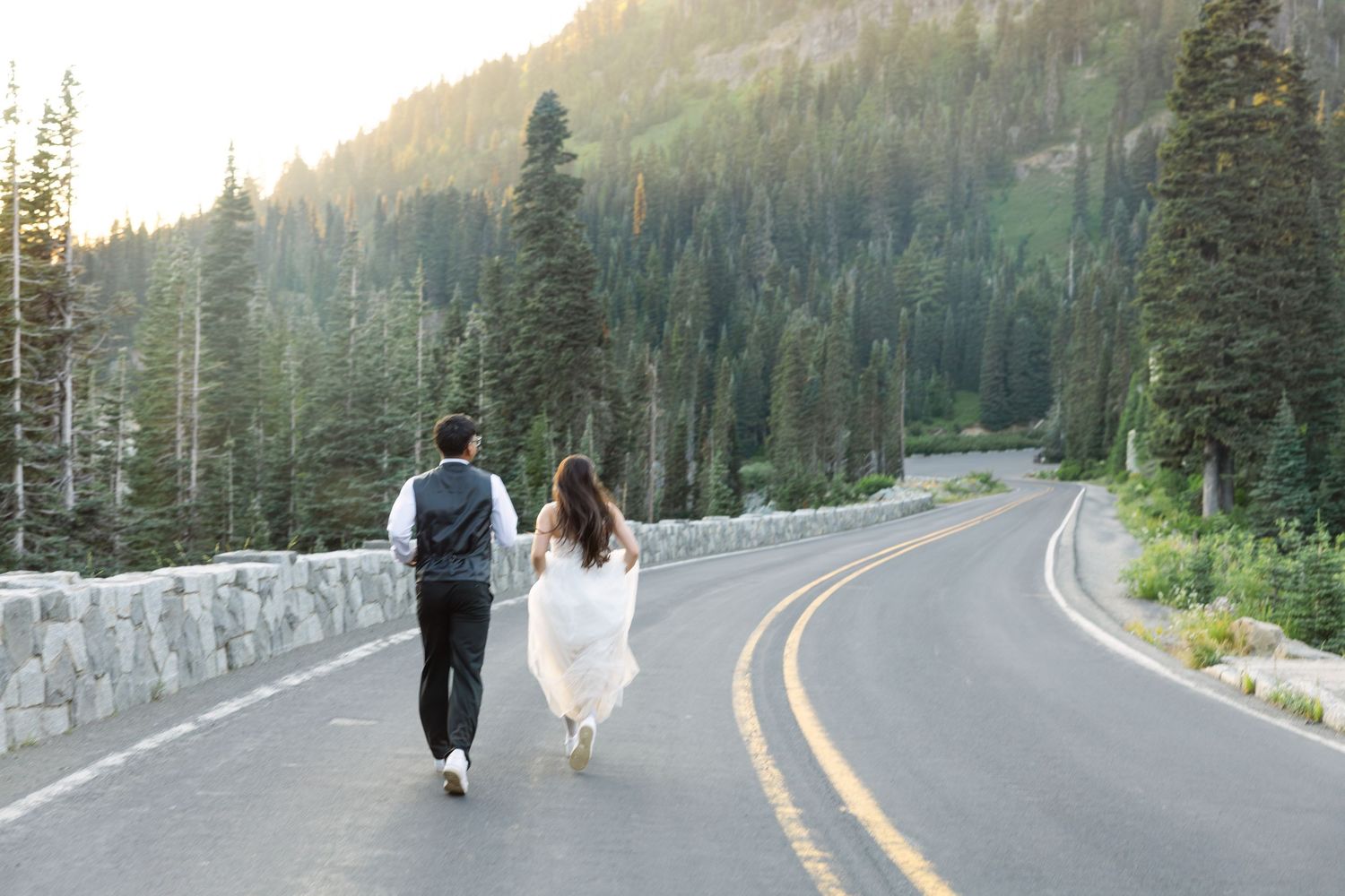 Two people walking down a scenic mountain road surrounded by evergreen trees and sunlit forest.