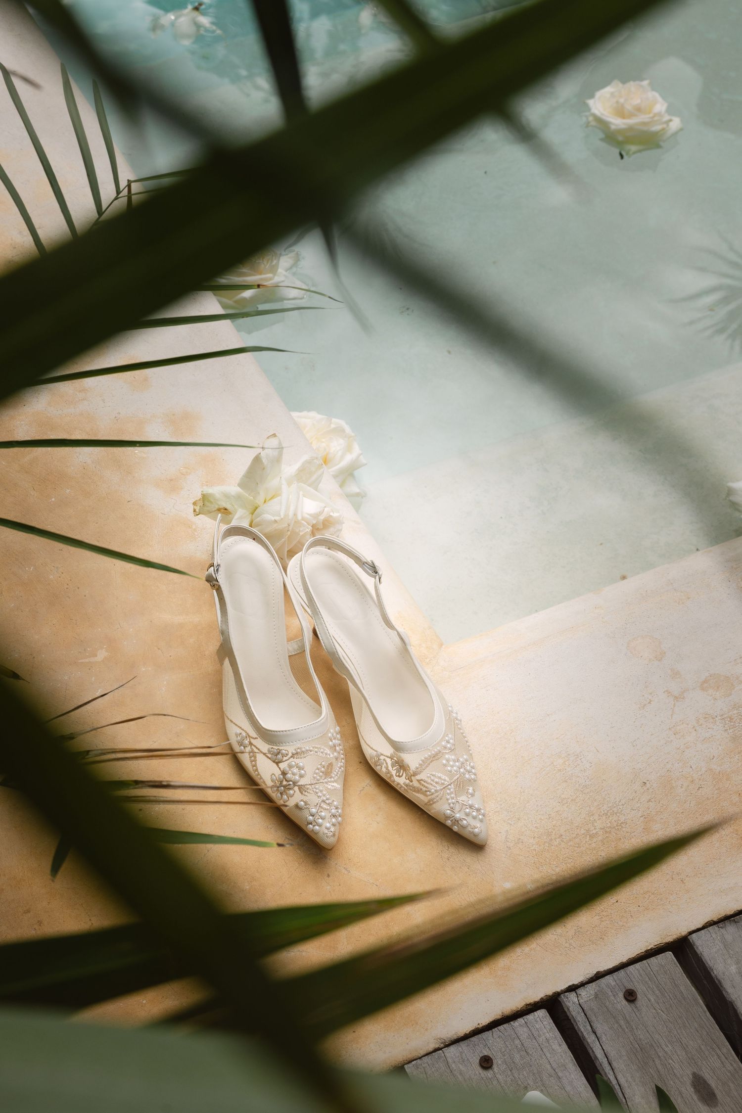 Elegant white bridal shoes with delicate lace details and beading sit on a light surface with palm leaf shadows.