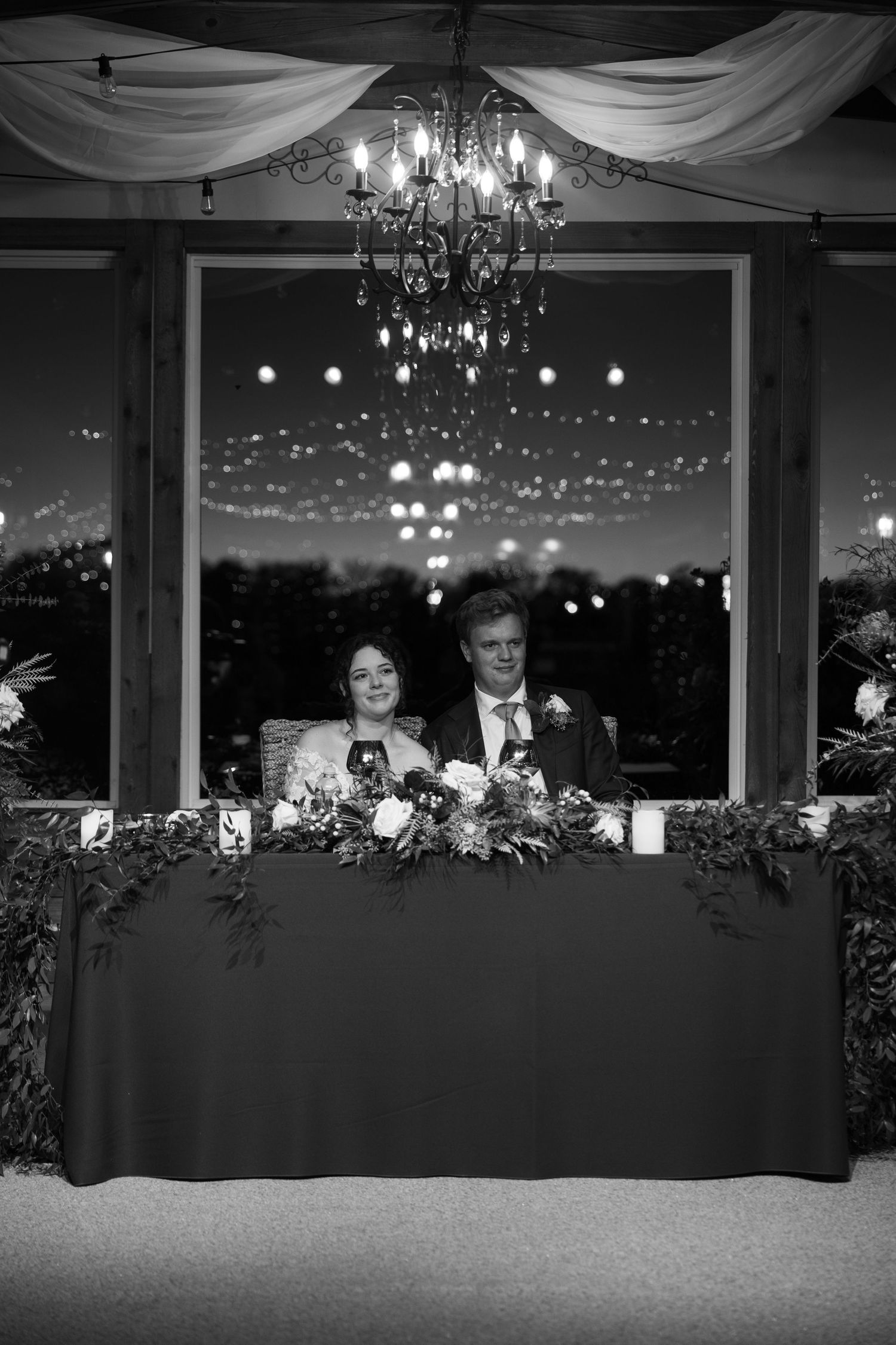 A black and white wedding reception photo showing a couple at their decorated head table beneath an elegant chandelier.