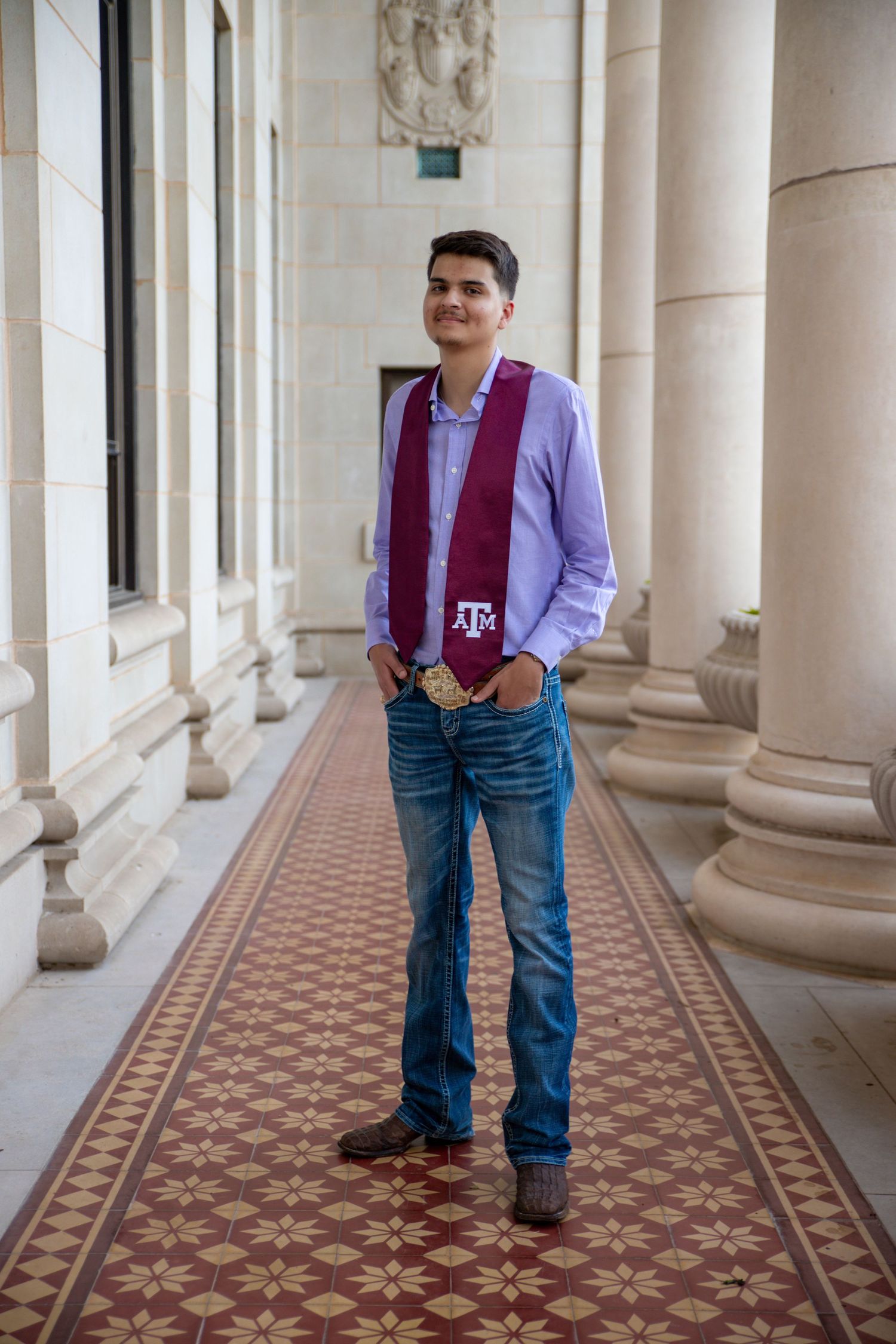 Student wearing light purple dress shirt and maroon Texas A&M scarf poses in classic campus hallway with ornate floor tiles.