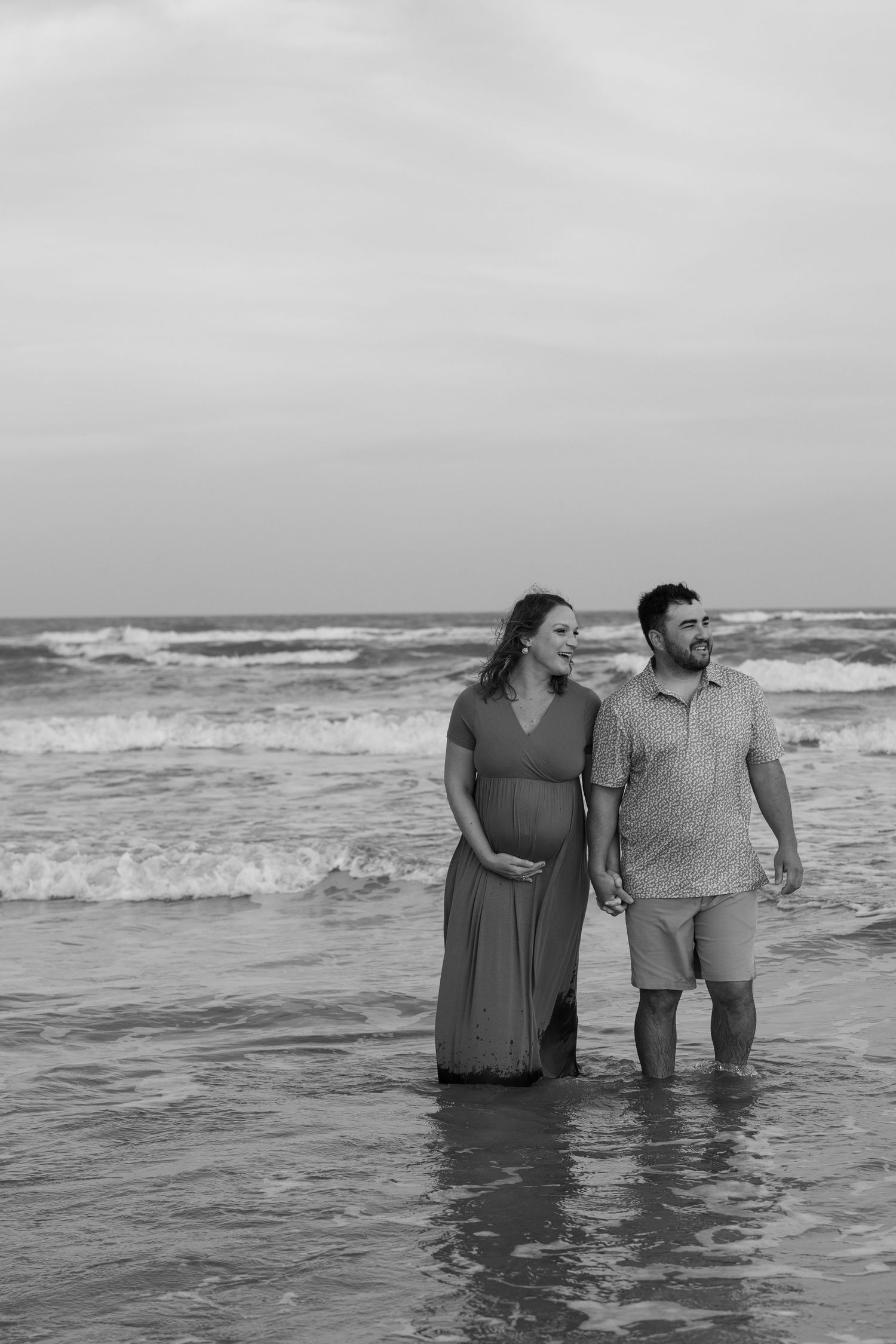 A couple stands together in ankle-deep ocean waves on a cloudy beach in black and white.