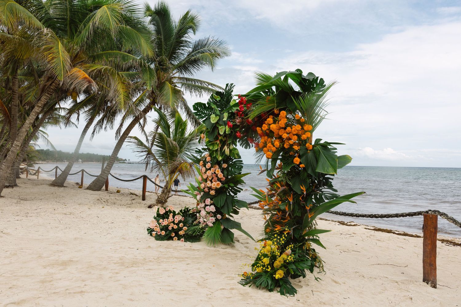 Palm trees line a tropical beach with wooden posts and orange flowers creating a scenic coastal pathway.