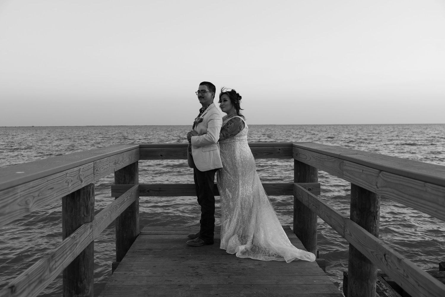A black and white photo of a couple standing back-to-back on a wooden pier overlooking the ocean.