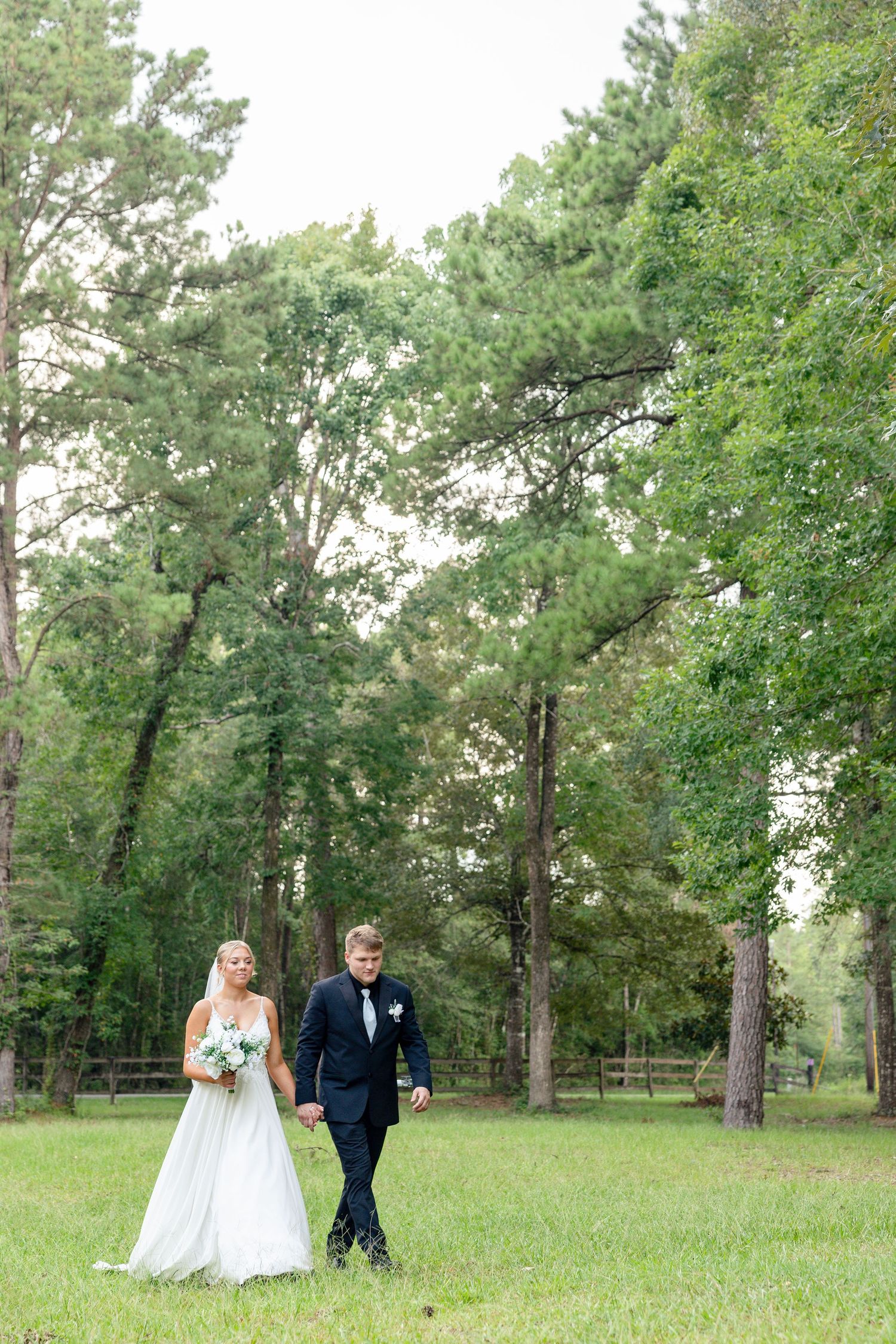 A couple walks together across a grassy lawn surrounded by tall green trees on their outdoor wedding day.