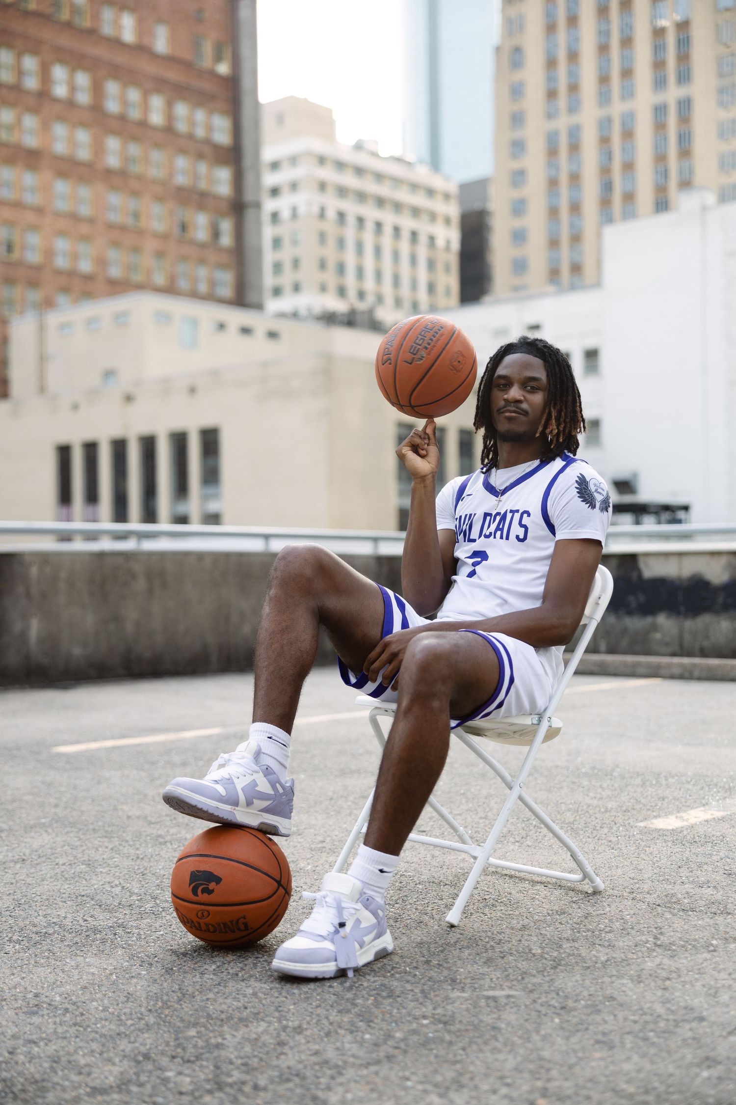 Basketball player in Kentucky Wildcats uniform sits in folding chair on urban rooftop with city buildings in background.