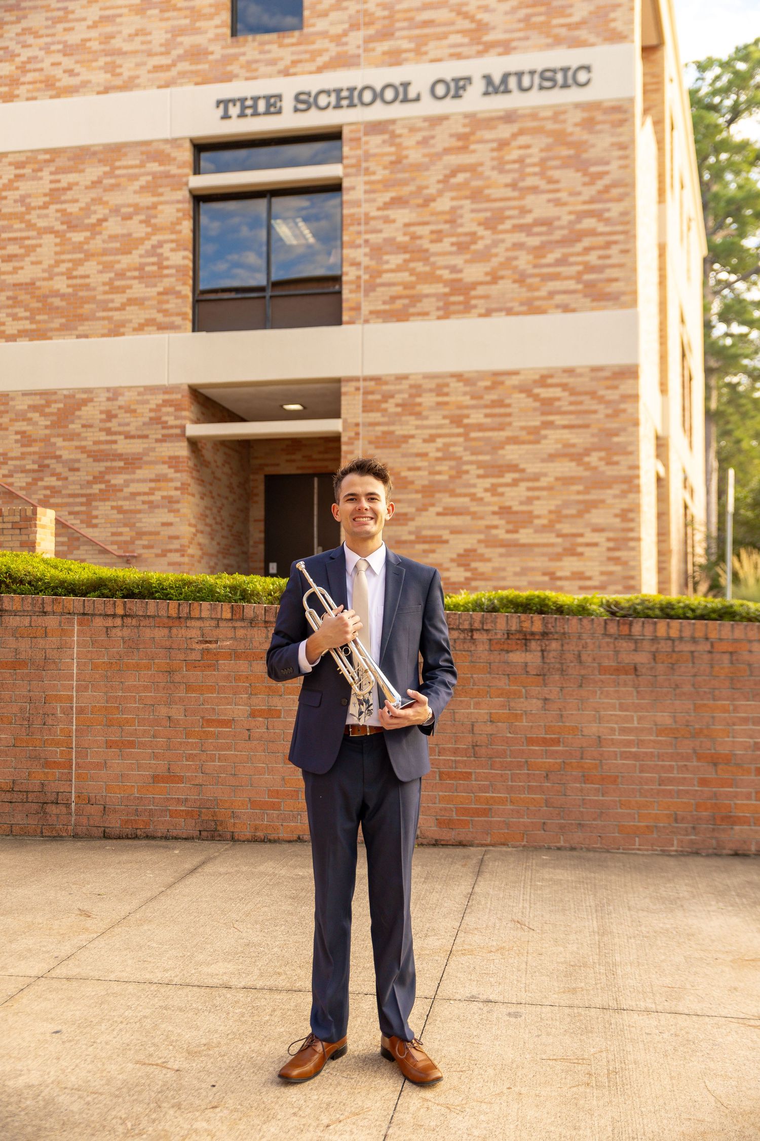 A musician in a navy suit holding a trumpet stands outside The School of Music building entrance.