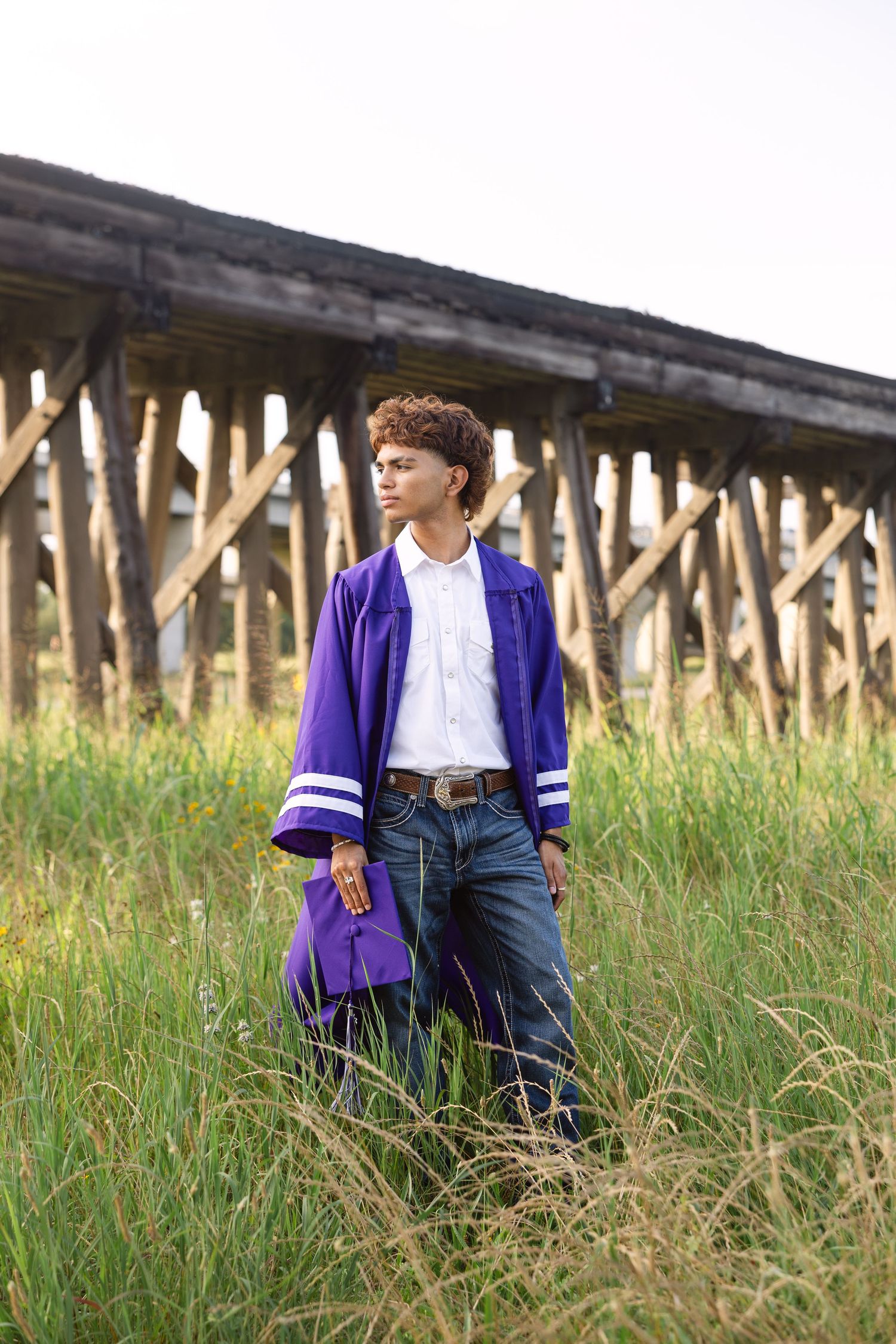 Someone wearing a purple varsity jacket and jeans stands in tall grass beneath a wooden railroad bridge structure.