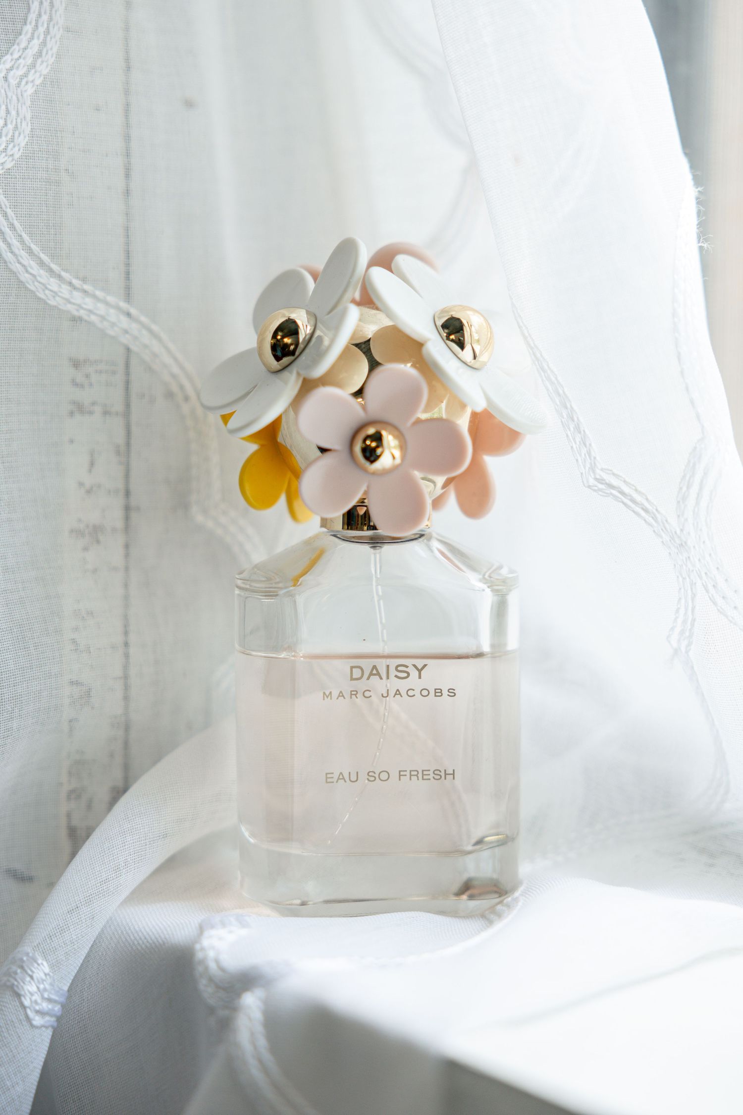 A bottle of Marc Jacobs Daisy Eau So Fresh perfume with delicate floral decorations on white fabric background.