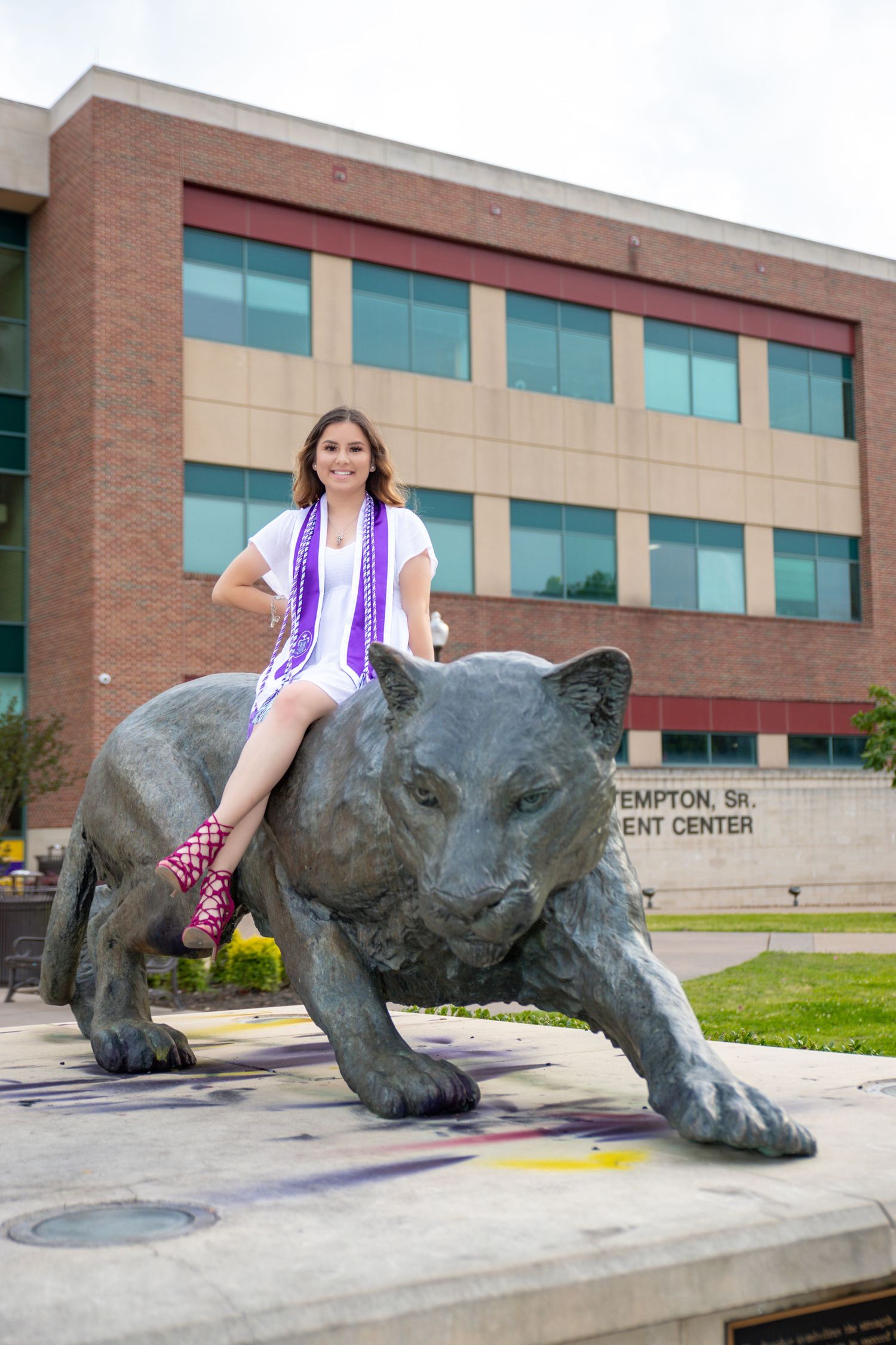 Graduate poses on large bronze panther statue outside the Hampton Student Center brick university building.