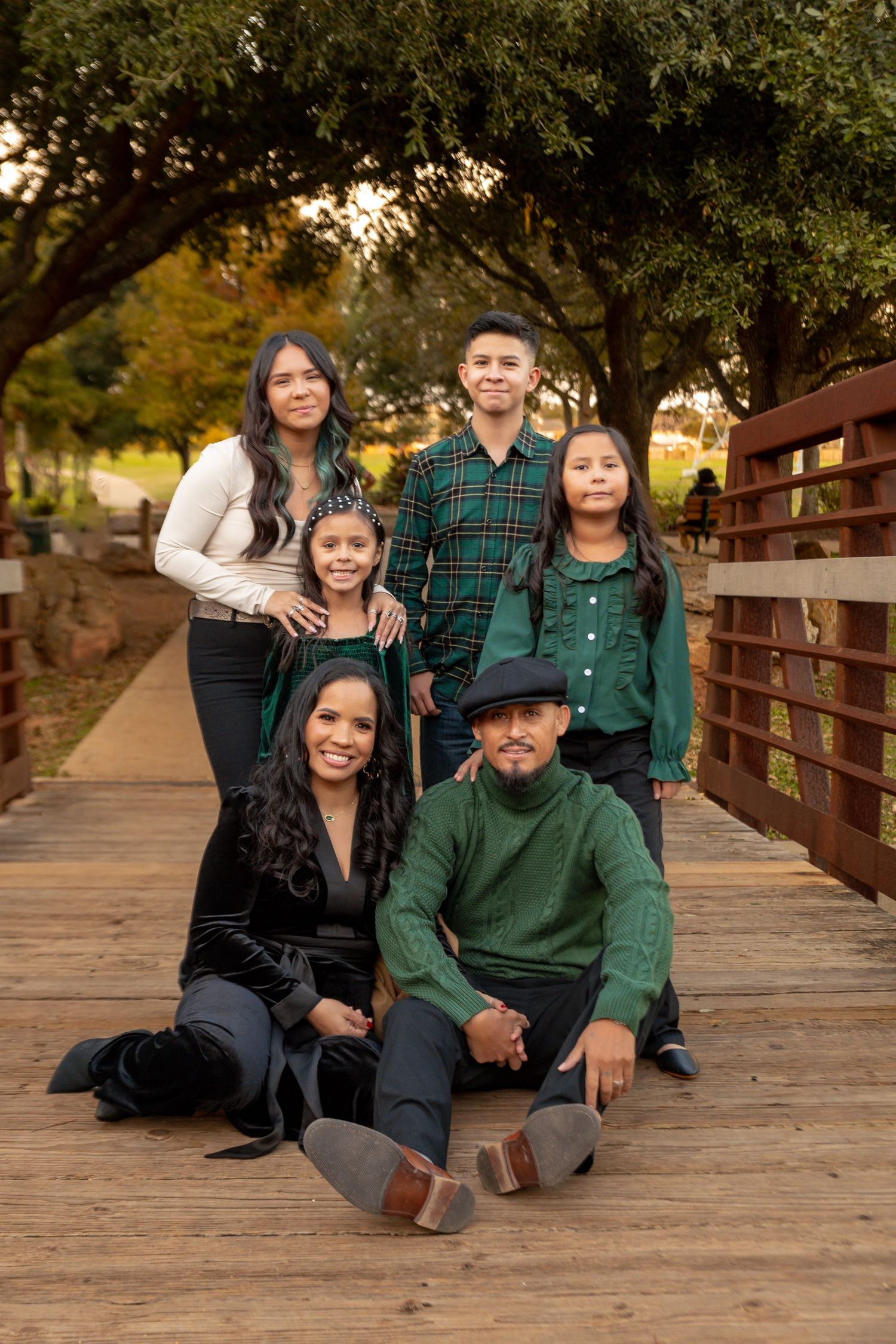Family wearing coordinated green and black outfits poses together on wooden bridge during outdoor photo session.