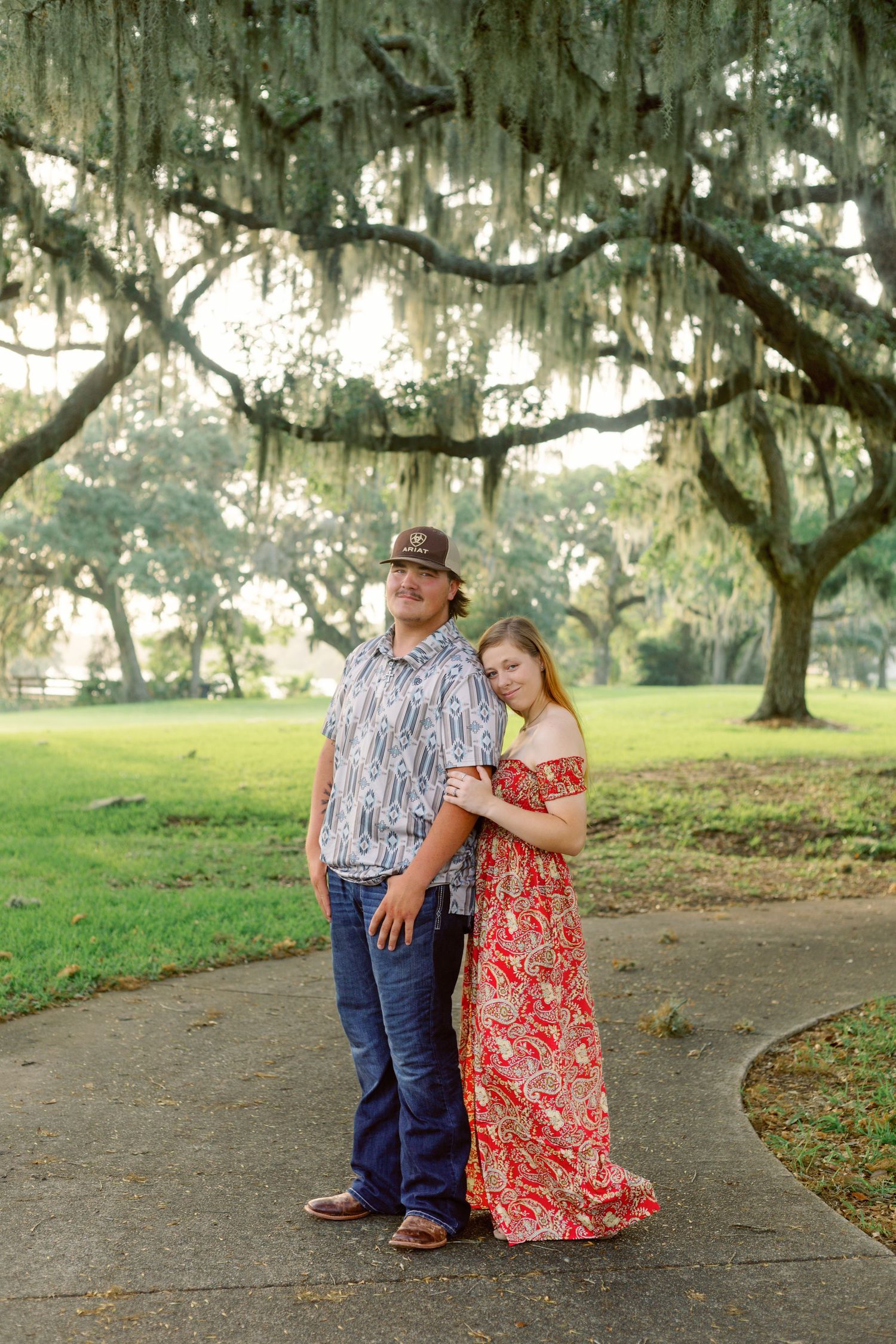 A couple shares a romantic moment under moss-draped oak trees in a Southern park setting.