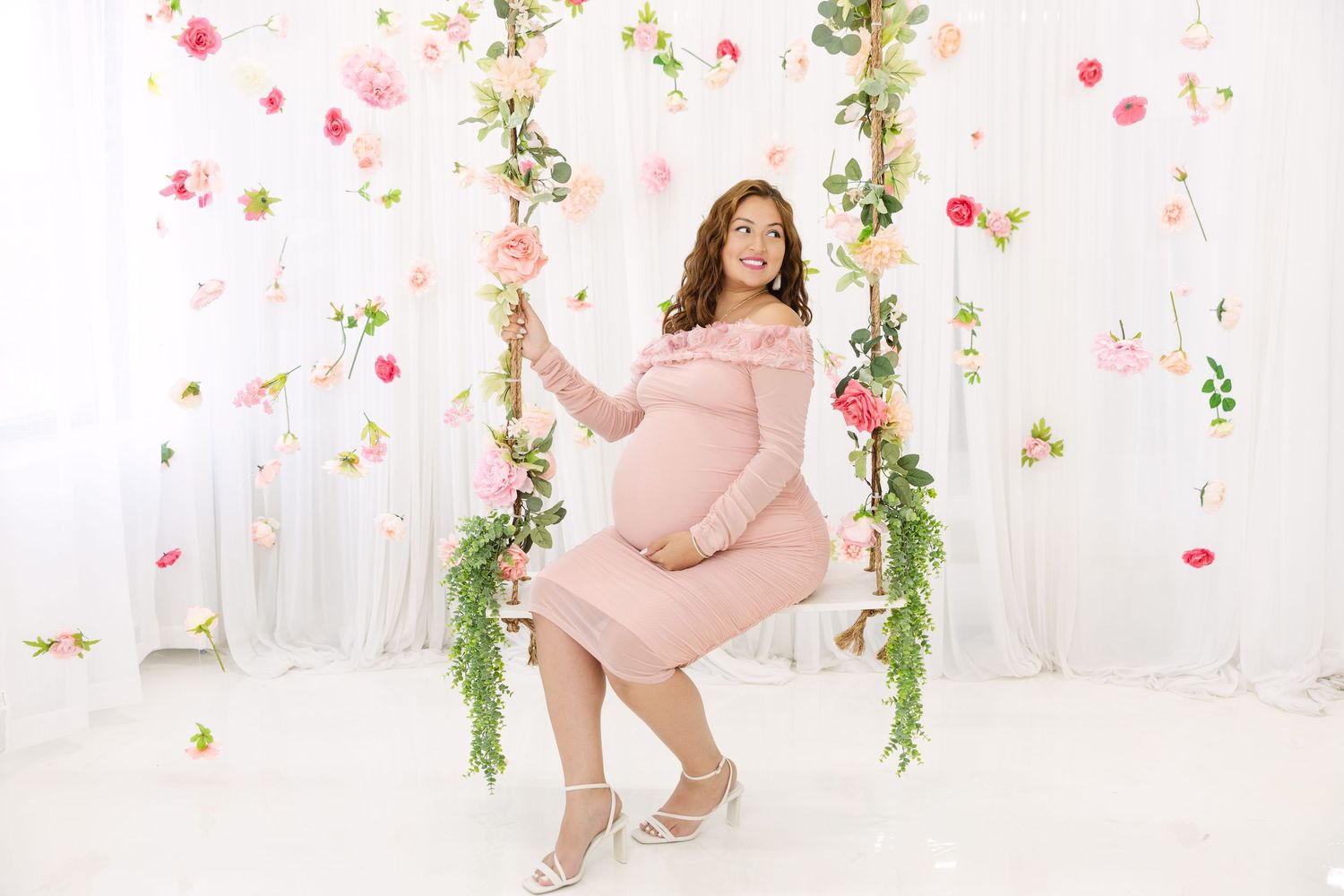 Someone wearing a pink dress poses on a decorative swing with floral arrangements and greenery against a white backdrop.