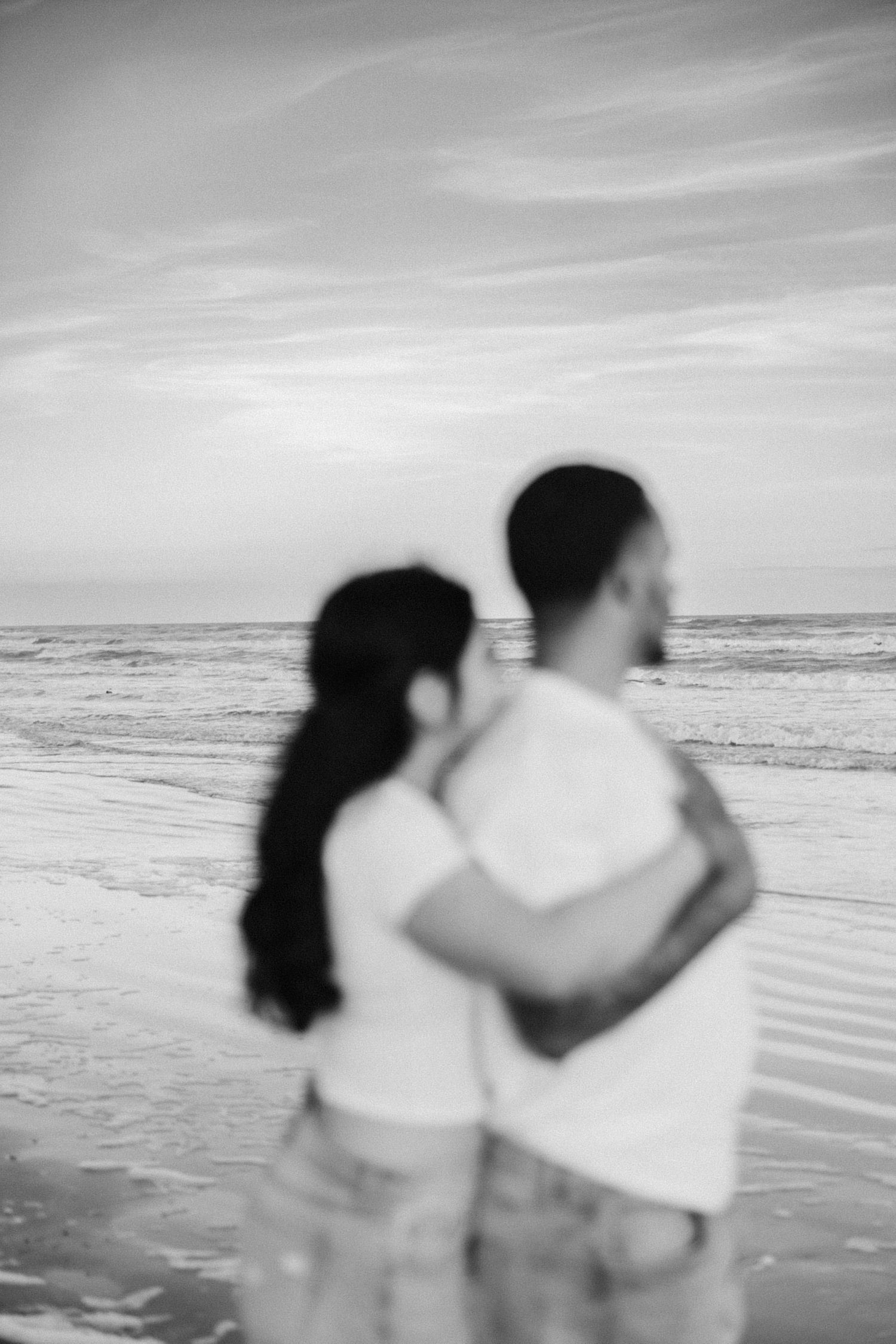 A couple stands close together looking out at the ocean waves in black and white photography on a beach.