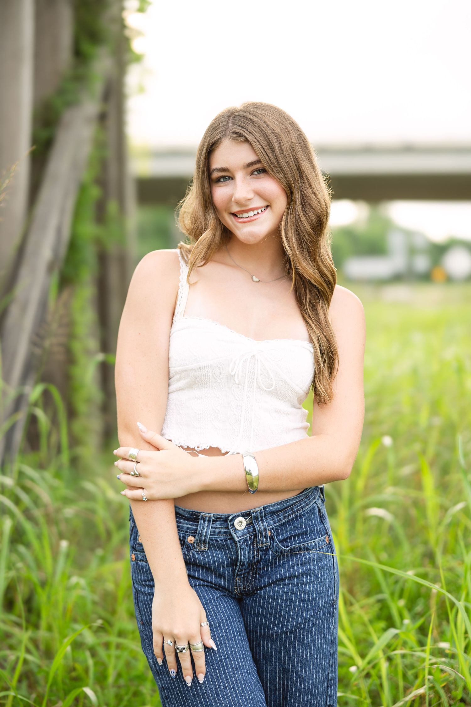 A person poses outdoors in a white crop top and jeans among tall grass on a sunny day.