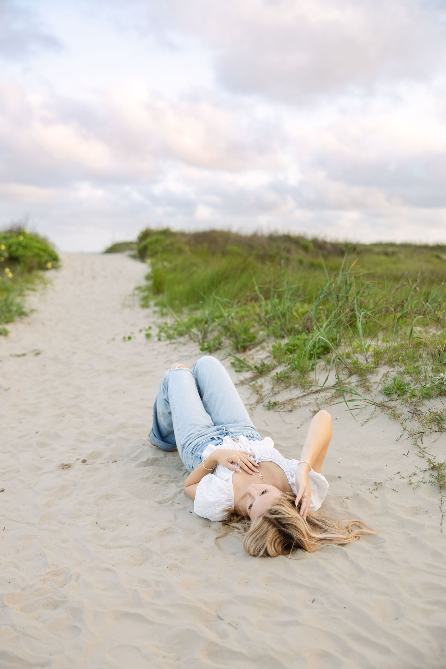 Person lying on sandy beach path with grass and dunes under cloudy sunset sky.