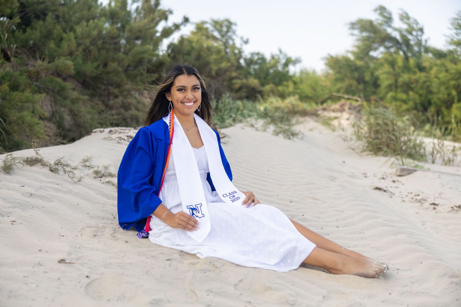 A person in a white dress and bright blue cardigan sits casually on a sandy beach path surrounded by vegetation.