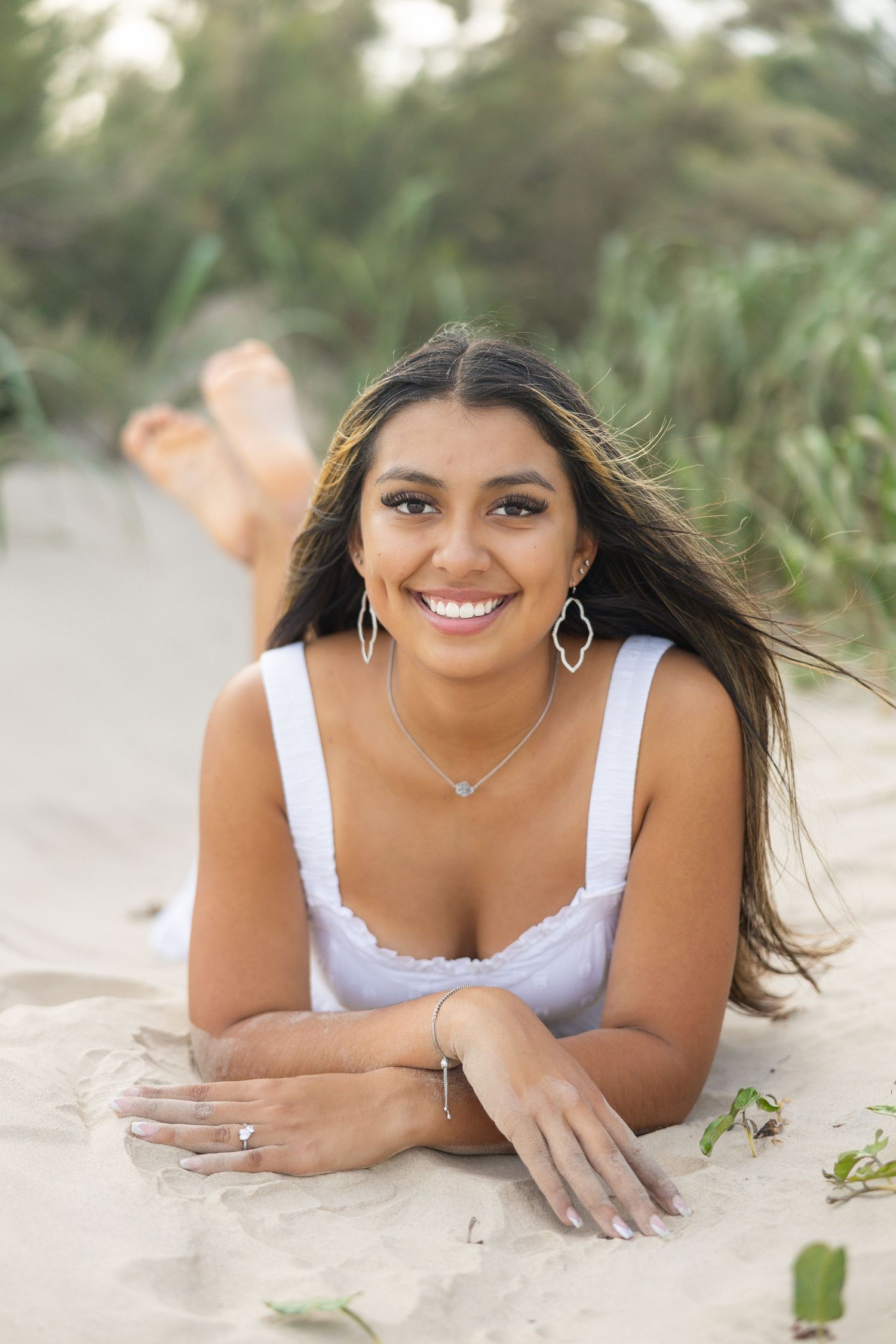 Smiling young woman in white top lying on sandy beach with windswept hair and silver jewelry.