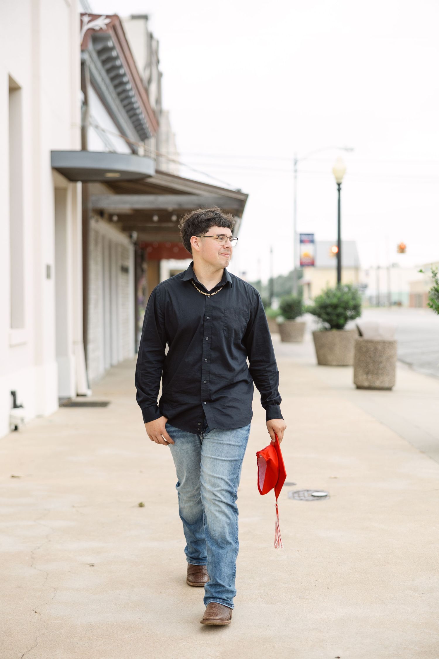 A person in a black button-down shirt and jeans walks down a sidewalk carrying a red item on a small-town street.