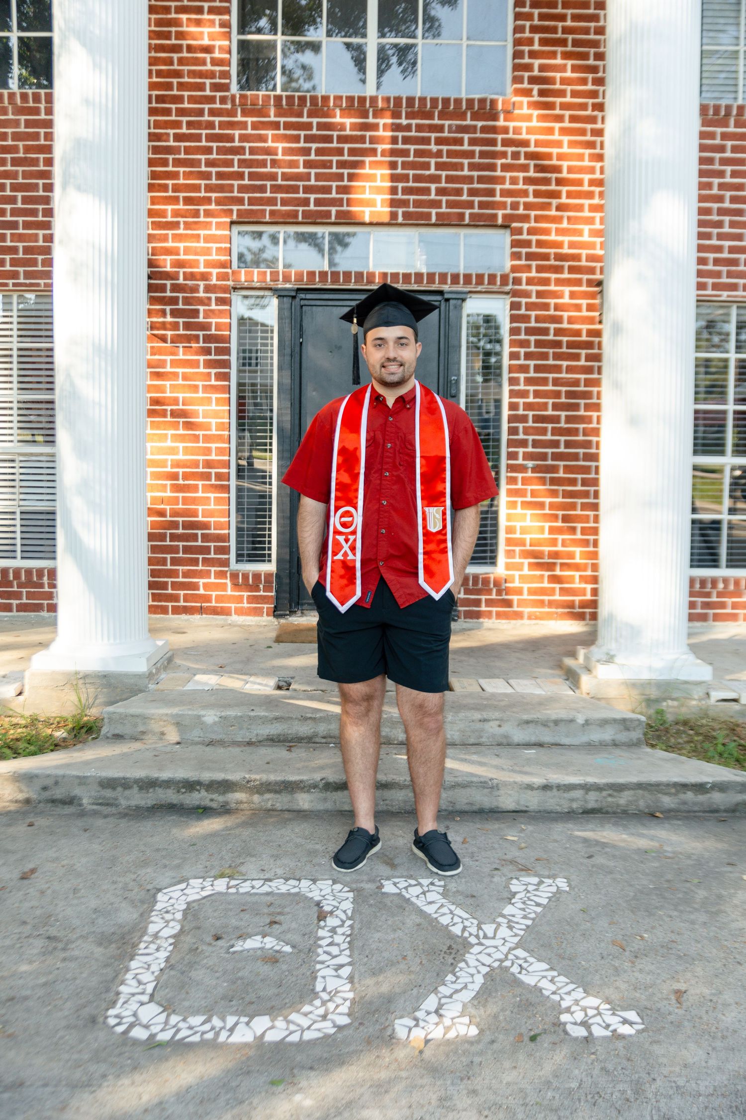 Graduate in cap and gown with red stoles stands in front of brick building with white columns and chalk arrows on ground.
