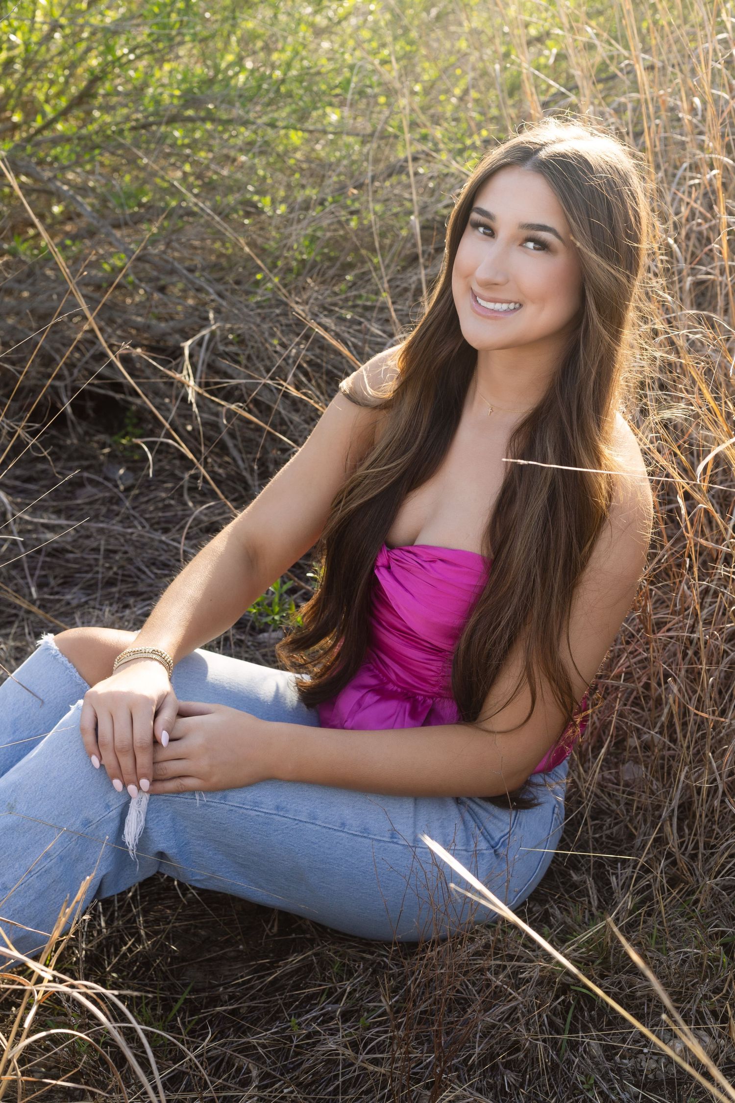 Young person in pink top and jeans sitting outdoors in tall grass during golden hour lighting.