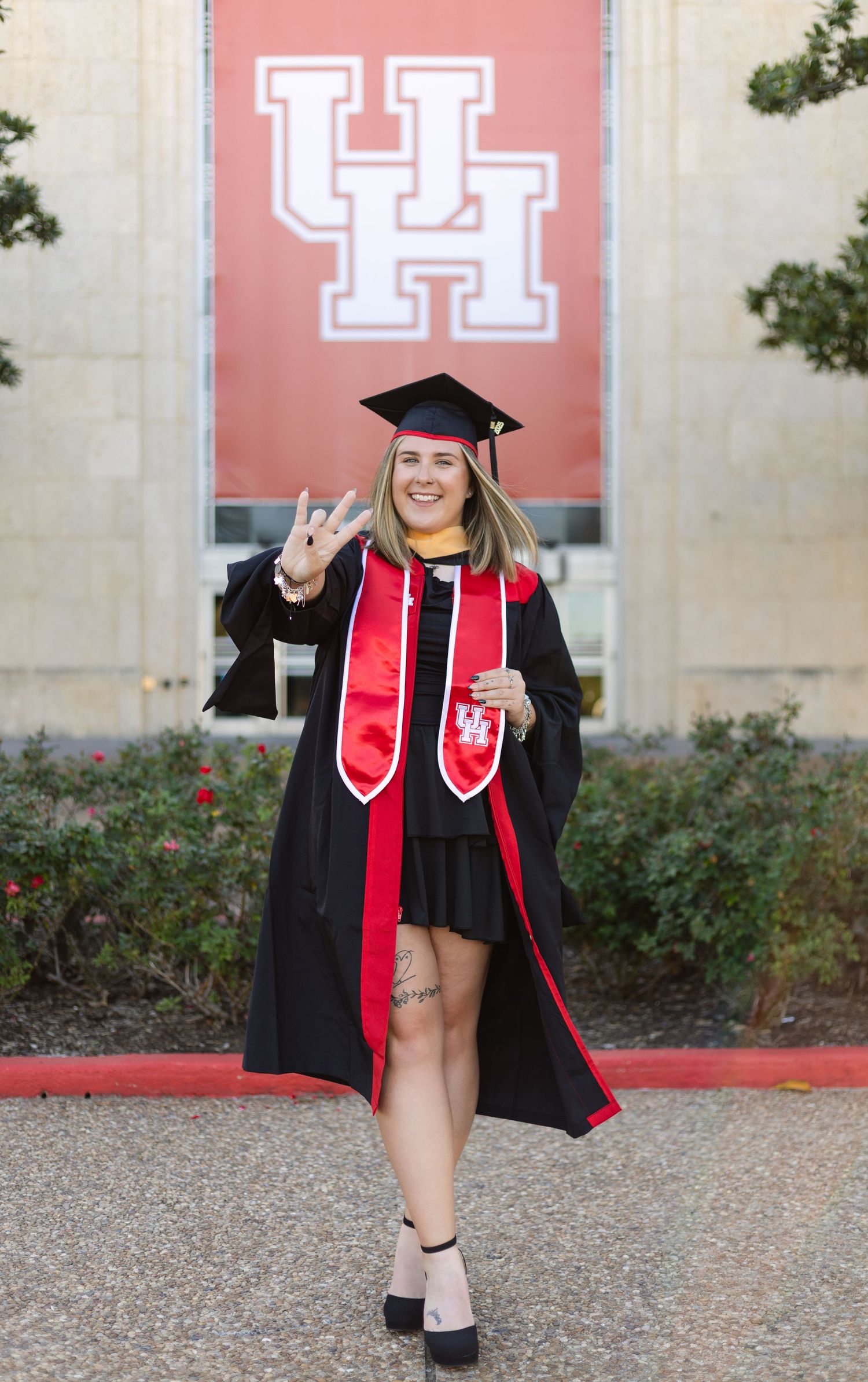 A graduate in cap and gown poses in front of a University of Houston logo during commencement.