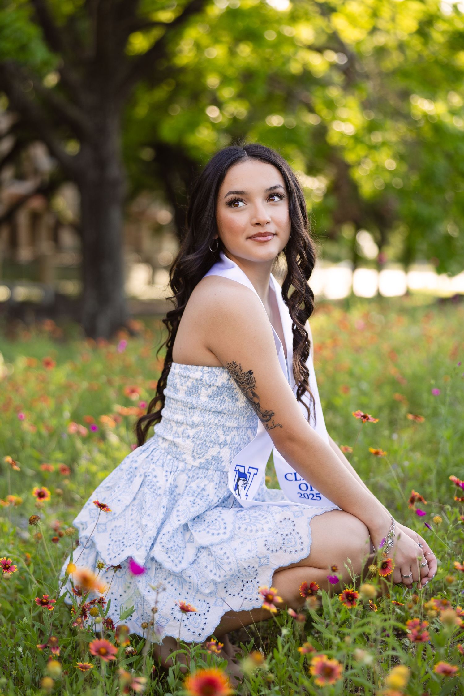A lovely portrait in a white lace dress sitting among wildflowers in a park during golden hour.