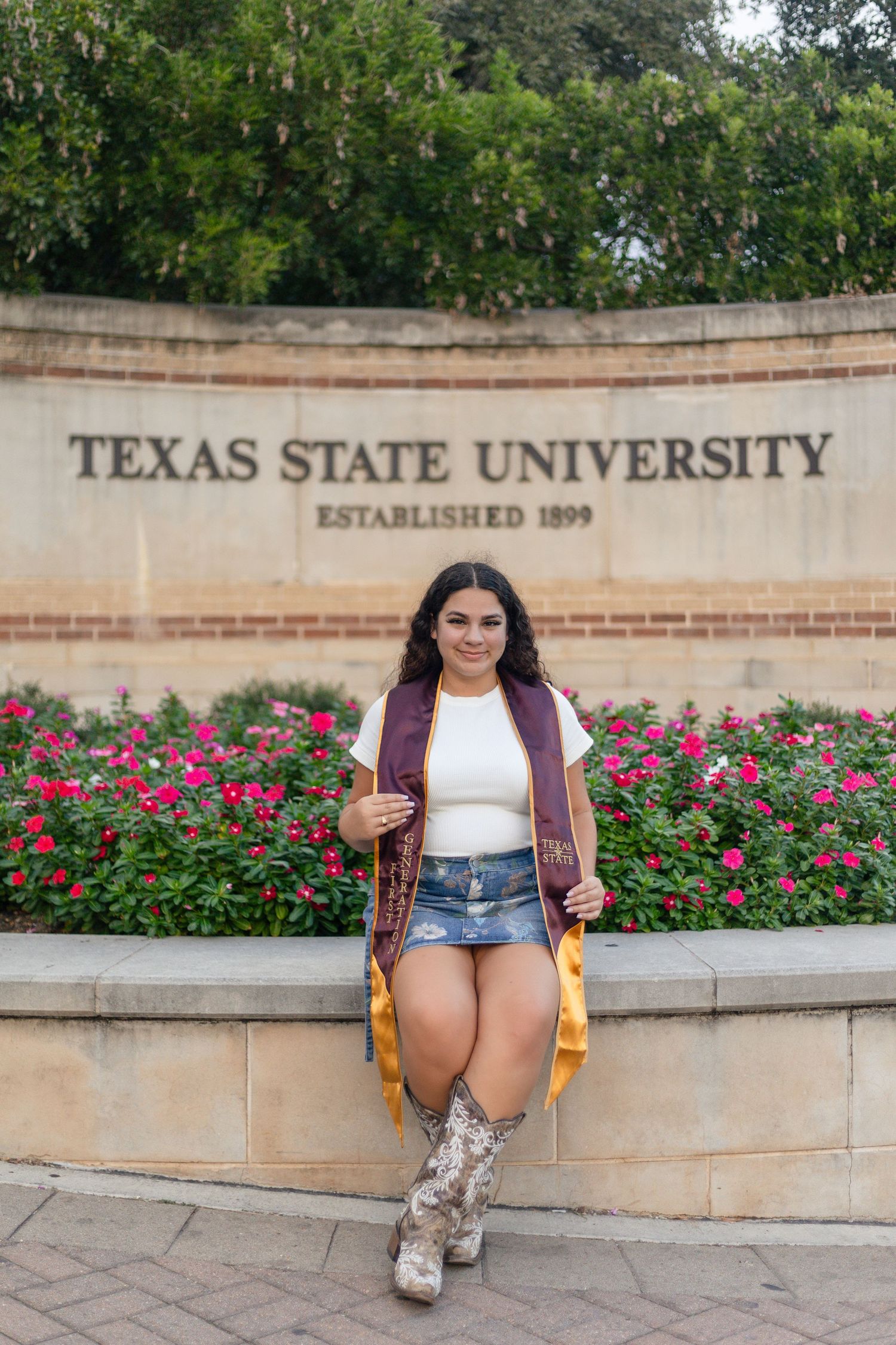 Student poses in front of Texas State University entrance sign with pink roses and brick wall backdrop.