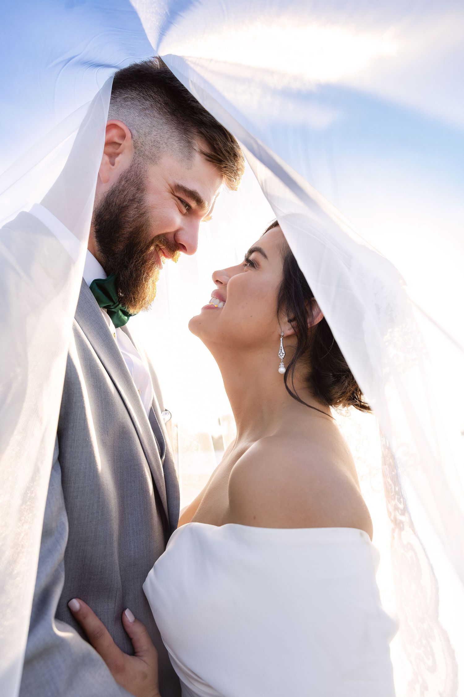 A romantic wedding photo of a couple sharing an intimate moment under a flowing veil against a bright sky.