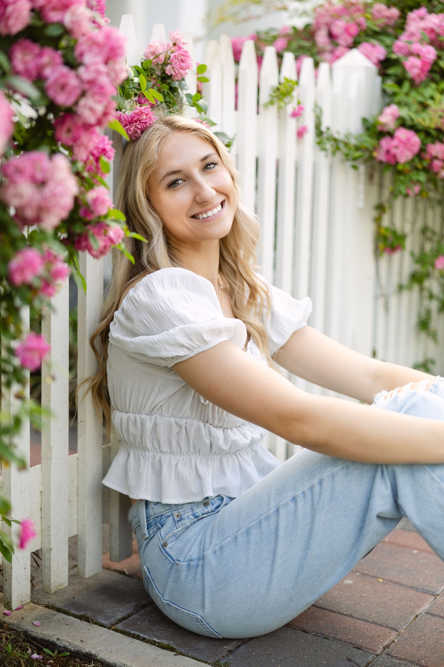 Person in white summer top and jeans sits by white picket fence with blooming pink roses on sunny day.