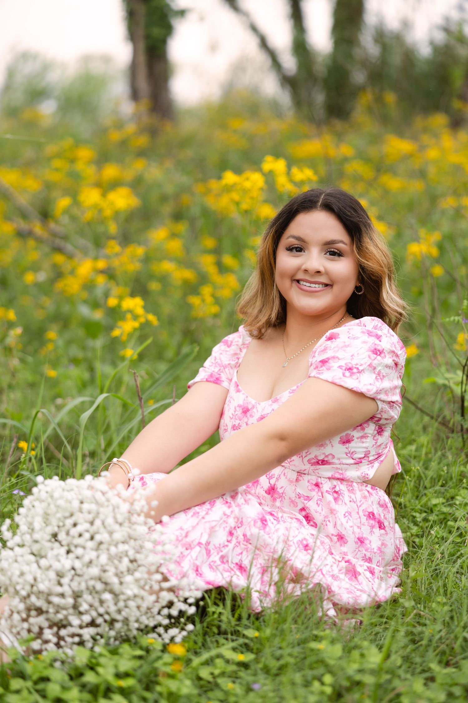 A figure in a pink floral dress sits in a meadow of yellow wildflowers holding white baby's breath bouquet.