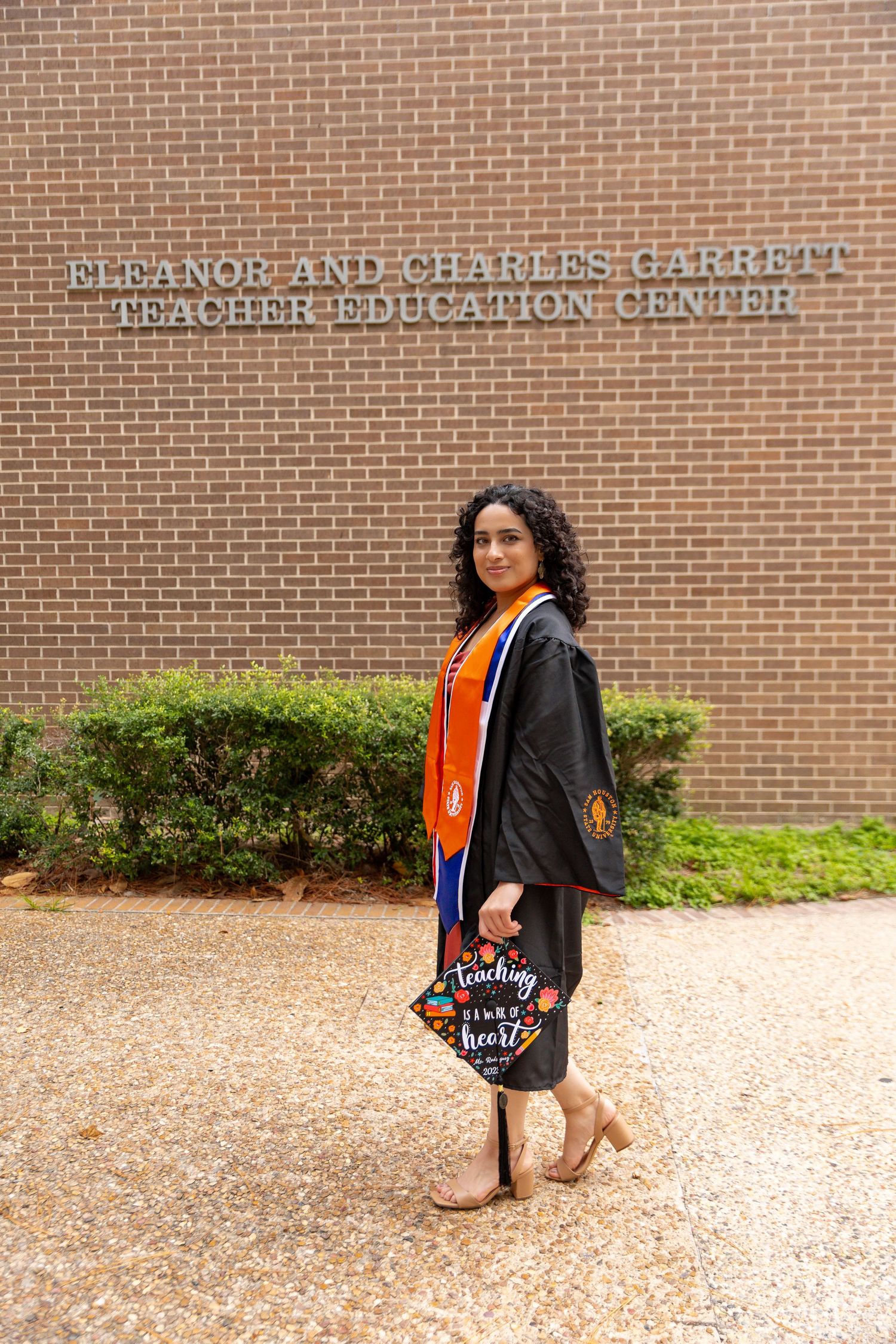 A graduate stands in a cap and gown outside the Eleanor and Charles U. Griffith Teacher Education Center building.