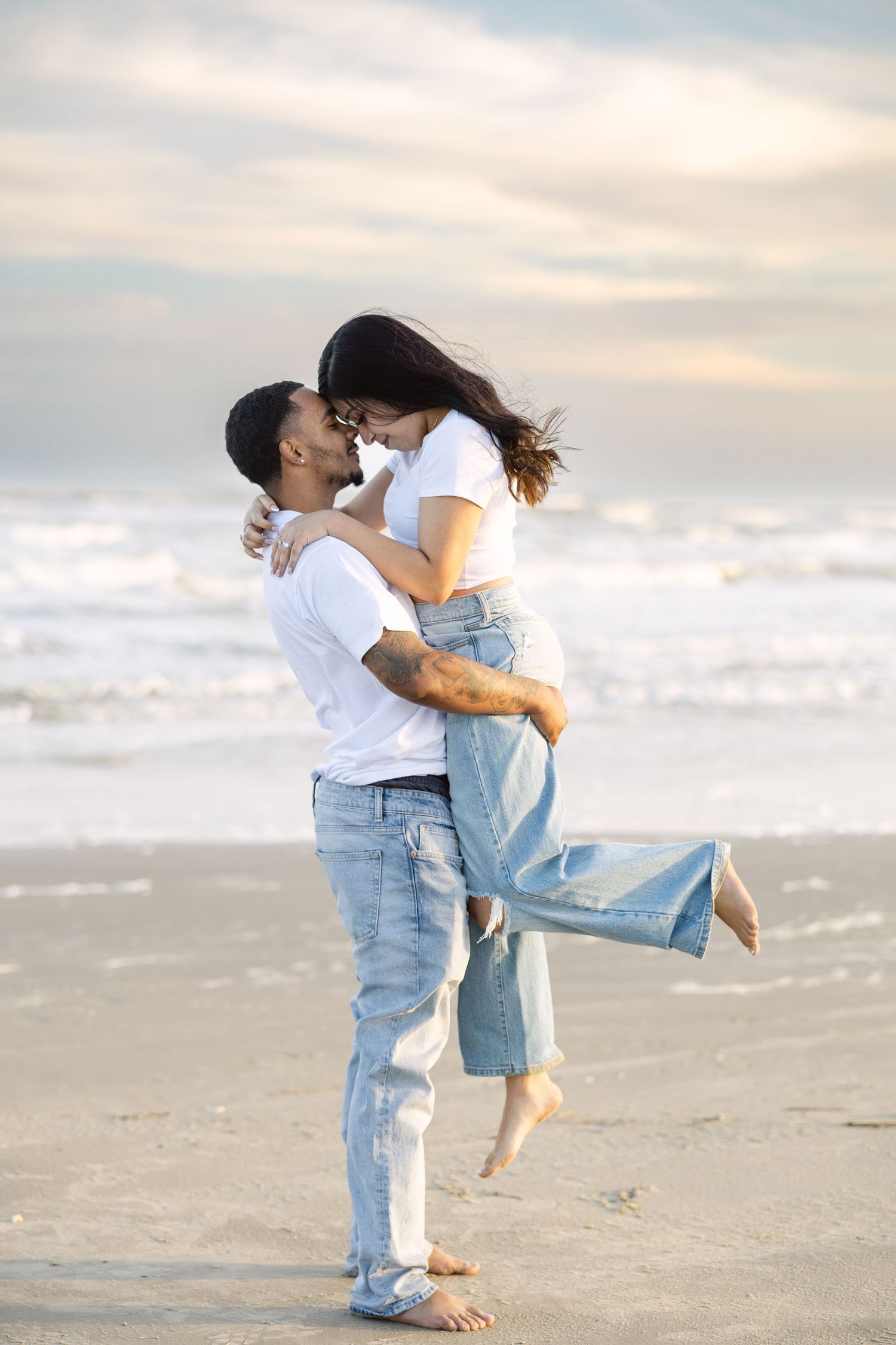 A romantic couple embracing and kissing on a misty beach at sunset while dressed in white tops and denim.