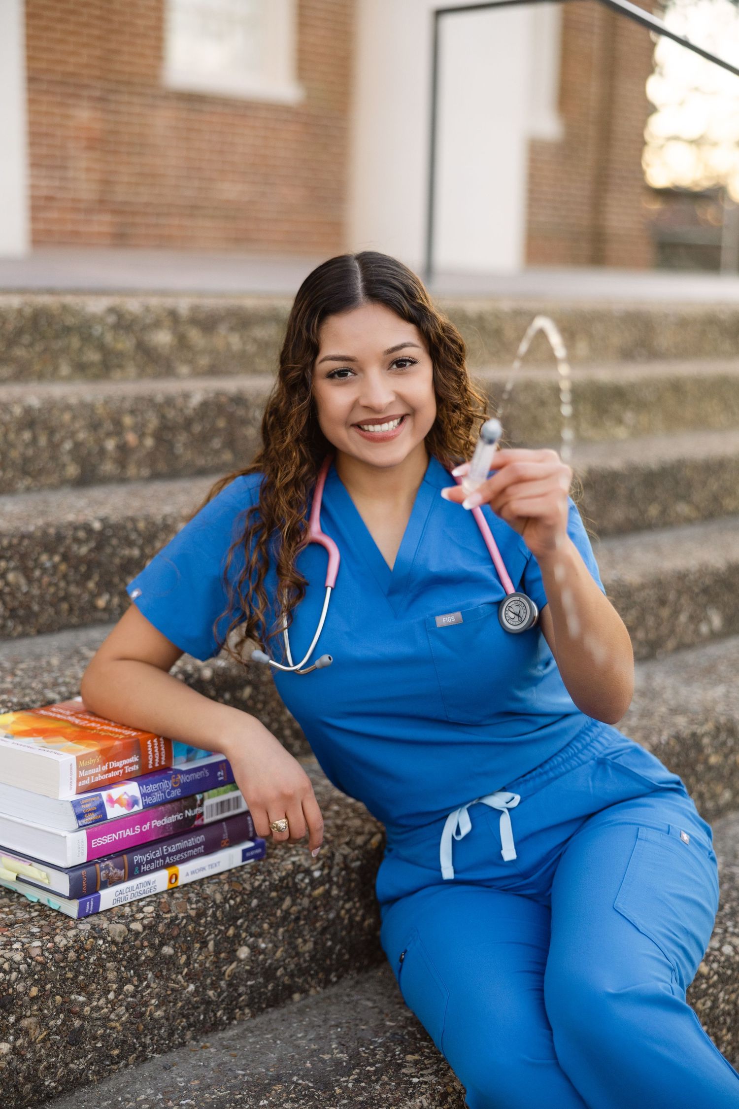 Healthcare professional in blue scrubs sitting on concrete steps with textbooks and medical equipment.