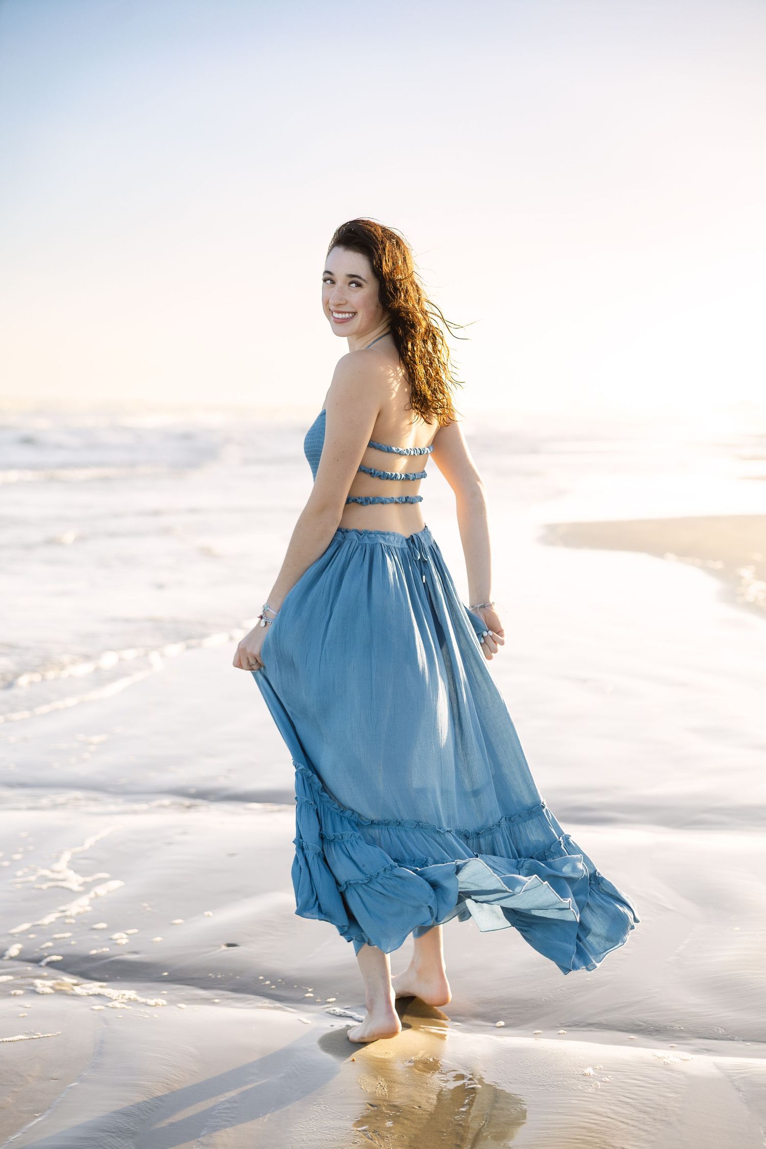 Flowing blue summer dress twirls on a sandy beach at sunset as ocean waves roll in along the shoreline.