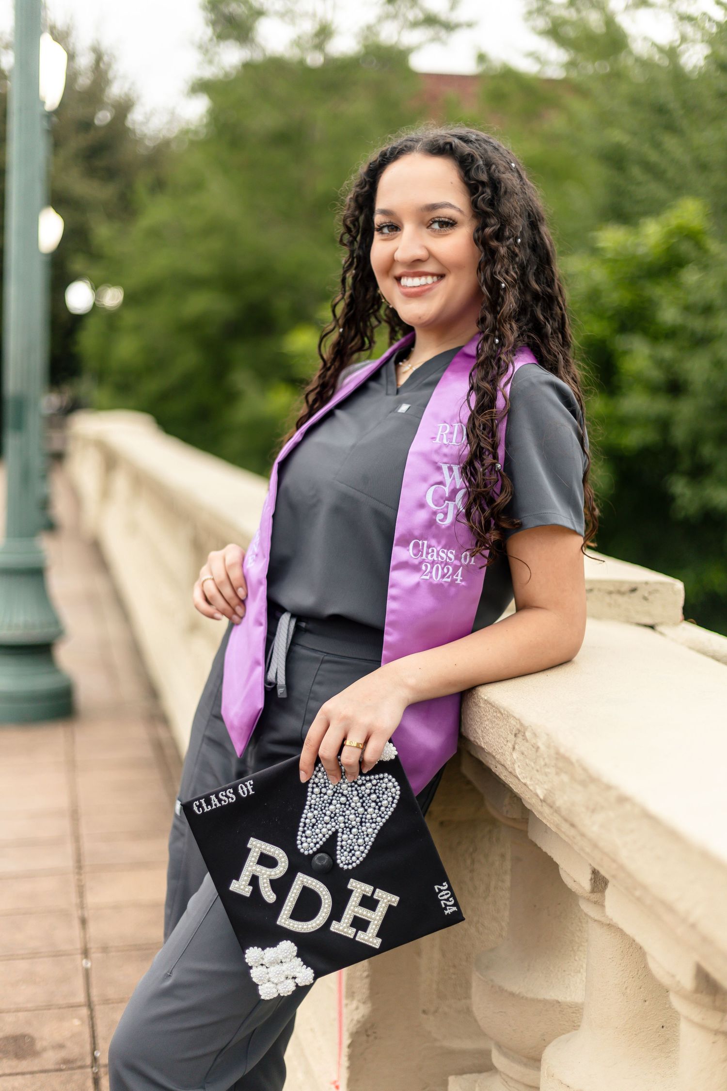 Graduate wearing black scrubs and purple graduation sash poses with decorated cap by stone railing outdoors.