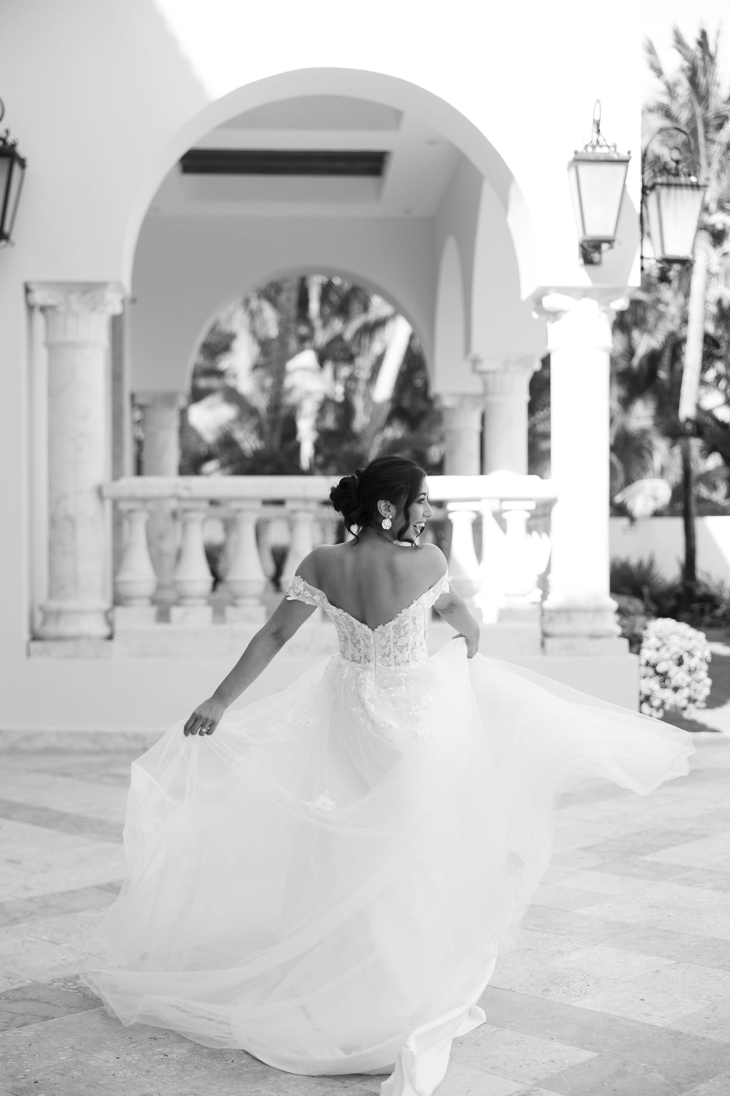 A bride in a flowing white wedding dress twirls gracefully under elegant white arches during a photo session.