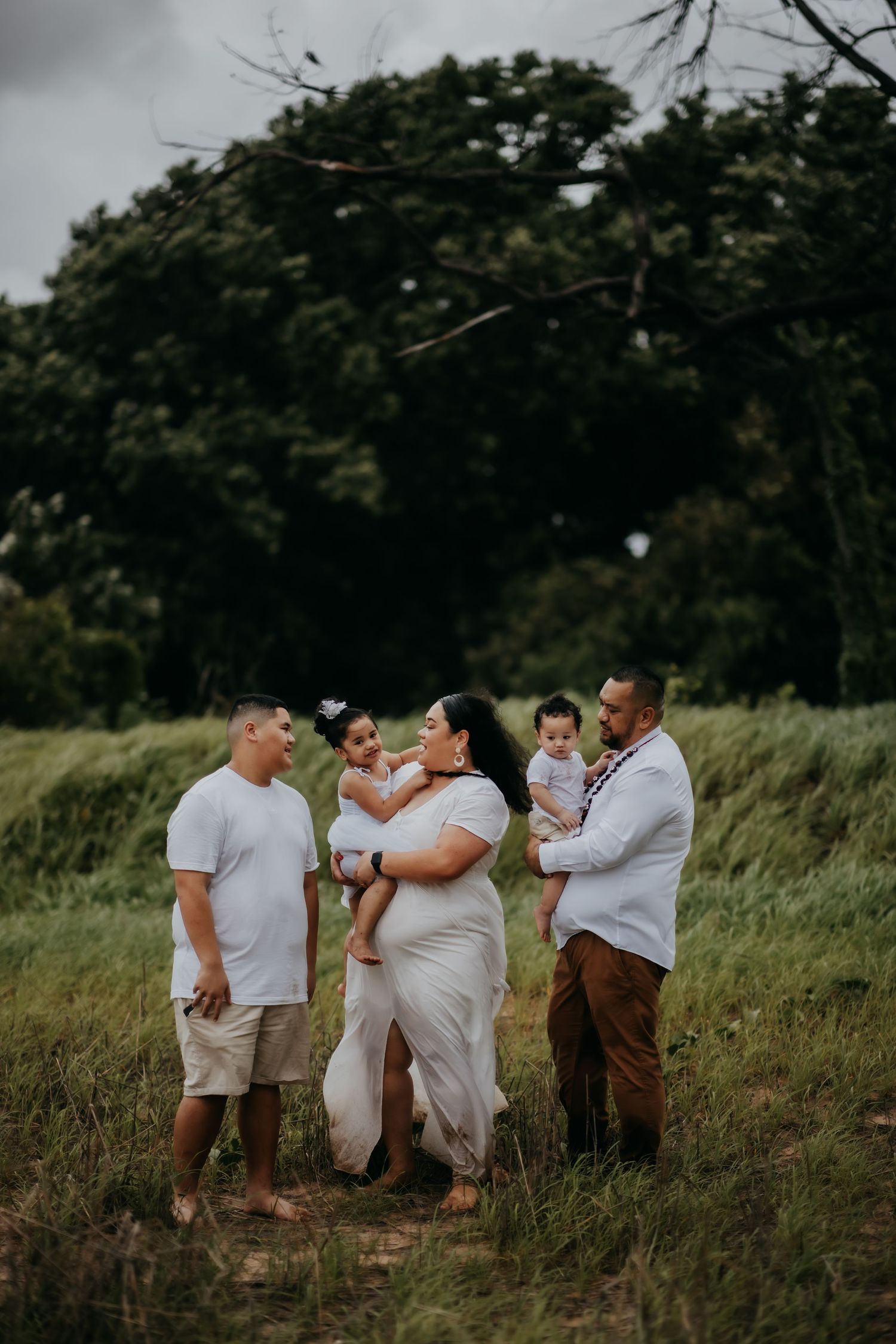 Family of five in white outfits standing in a lush green field, parents holding two young children.