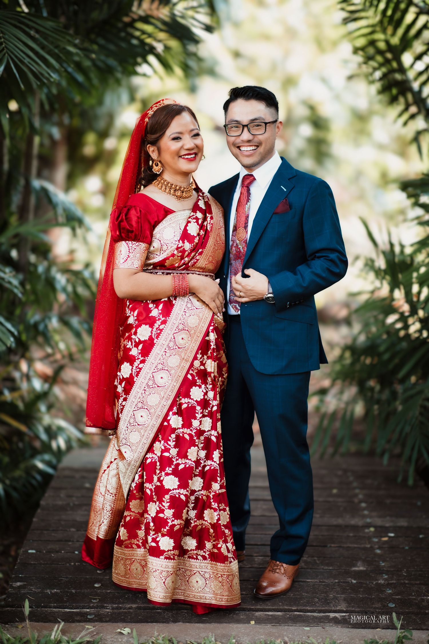 Indian bride in red floral saree with groom in navy suit posing together in lush garden setting.