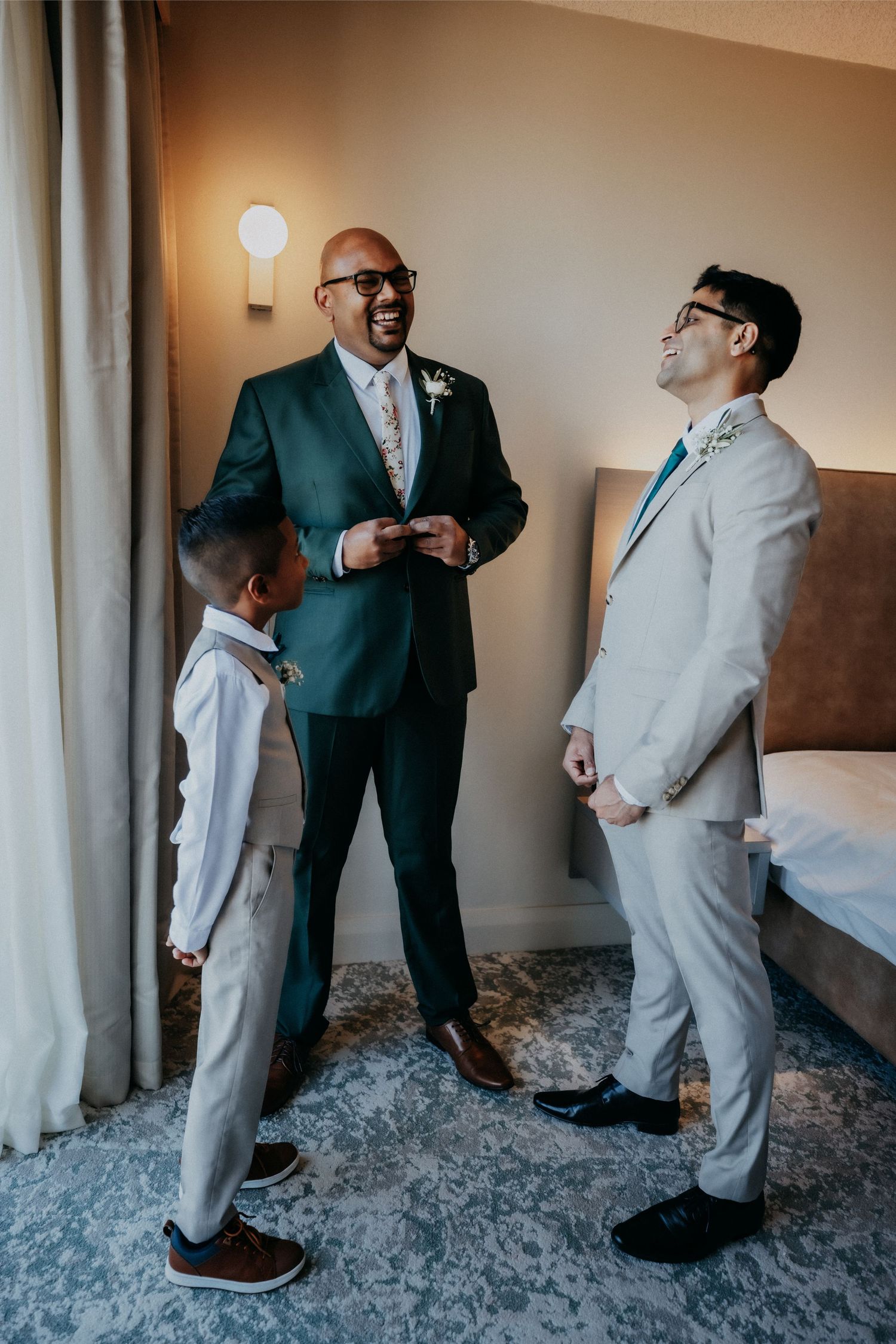 Groom in green suit laughing with groomsman and young boy in hotel room before wedding ceremony.