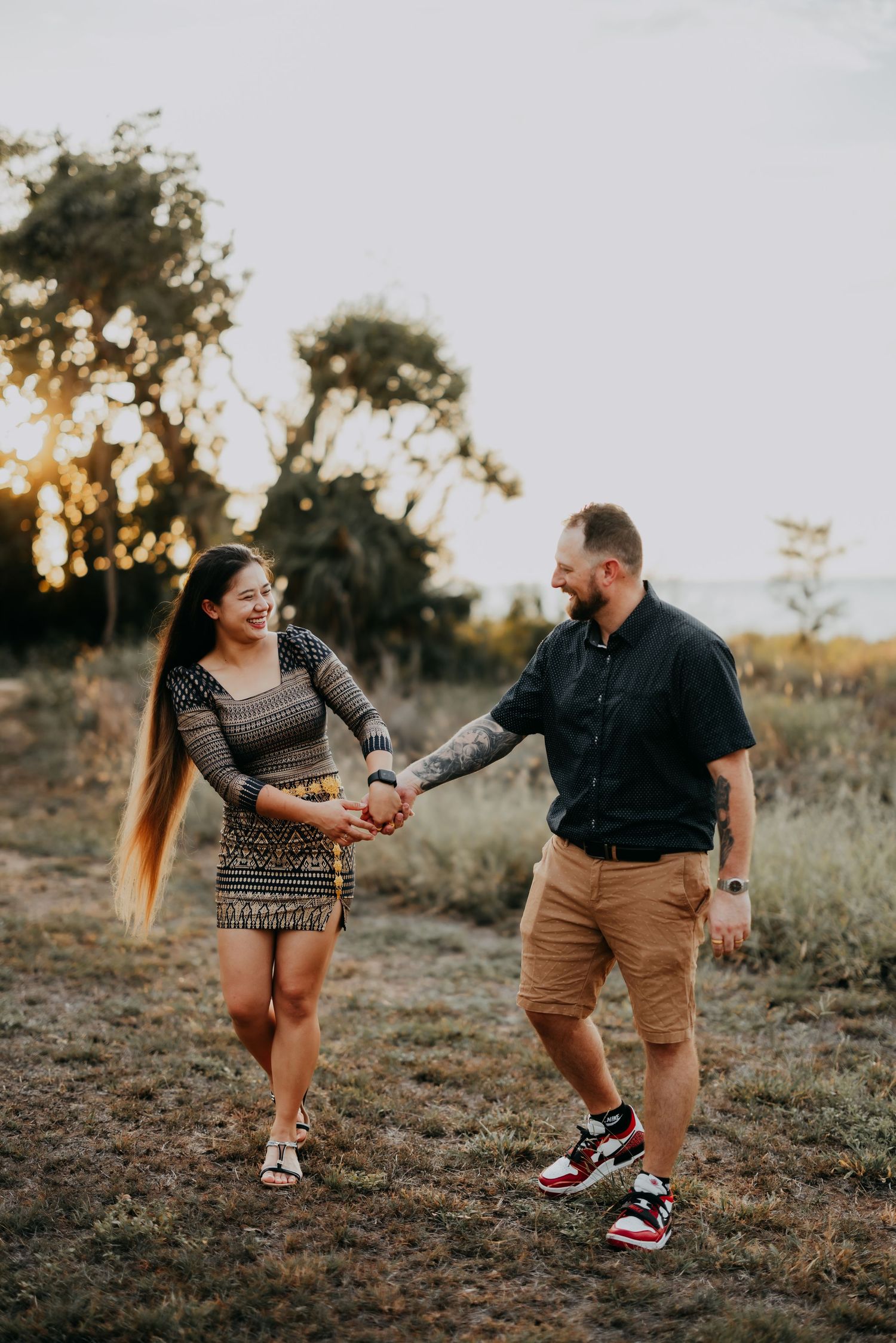 Couple holding hands and laughing while walking through a sunlit field at sunset during golden hour.