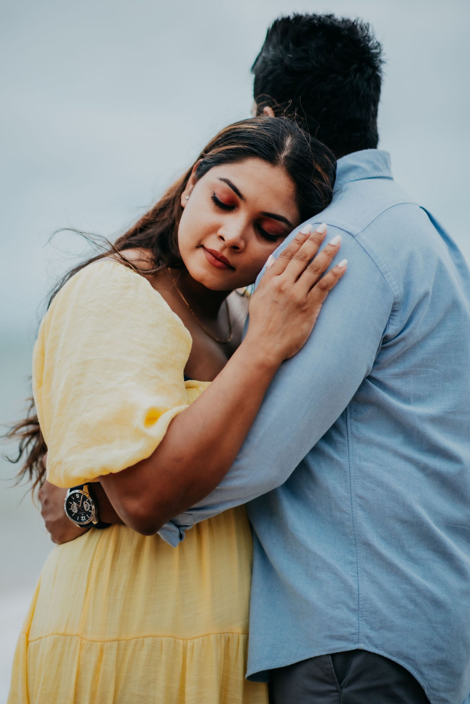 Woman in yellow dress tenderly embracing man in blue shirt during emotional beach photo session.